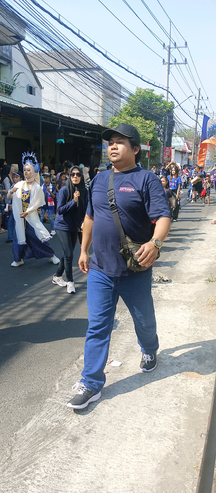 In Malang, Indonesia, a man in a dark blue t-shirt, blue jeans, and a black baseball cap walks on a sunlit street. He wears a gold watch and a green crossbody bag. Behind him, a woman in a dark blue hijab and sunglasses walks, her hands clasped. To her left, a woman in traditional attire, including a blue and white dress and an elaborate silver headdress, also walks. Other people, some in blue t-shirts, are visible further down the street. The scene appears to be a street procession or parade, with buildings lining the right side of the road and electrical wires overhead.