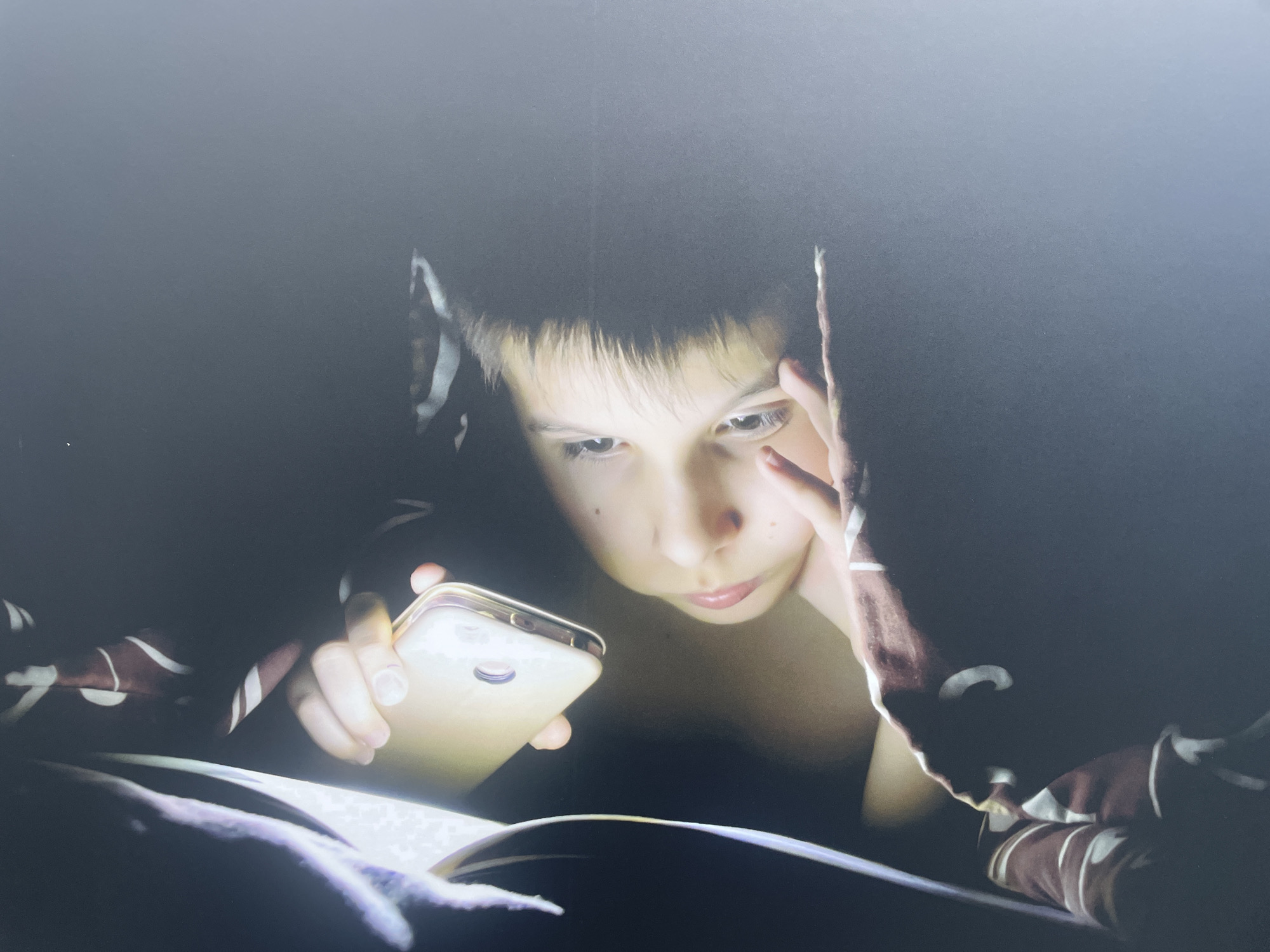 A person is illuminated by the light of a mobile phone screen while lying under bedcovers patterned with white and brown stripes. One hand holds the phone, while the other is near their face, possibly resting on their forehead. The surrounding environment is dark, suggesting it is night, and the scene is likely indoors, possibly a bedroom. There is no information in the image indicating the location is Sochi, Russia.
