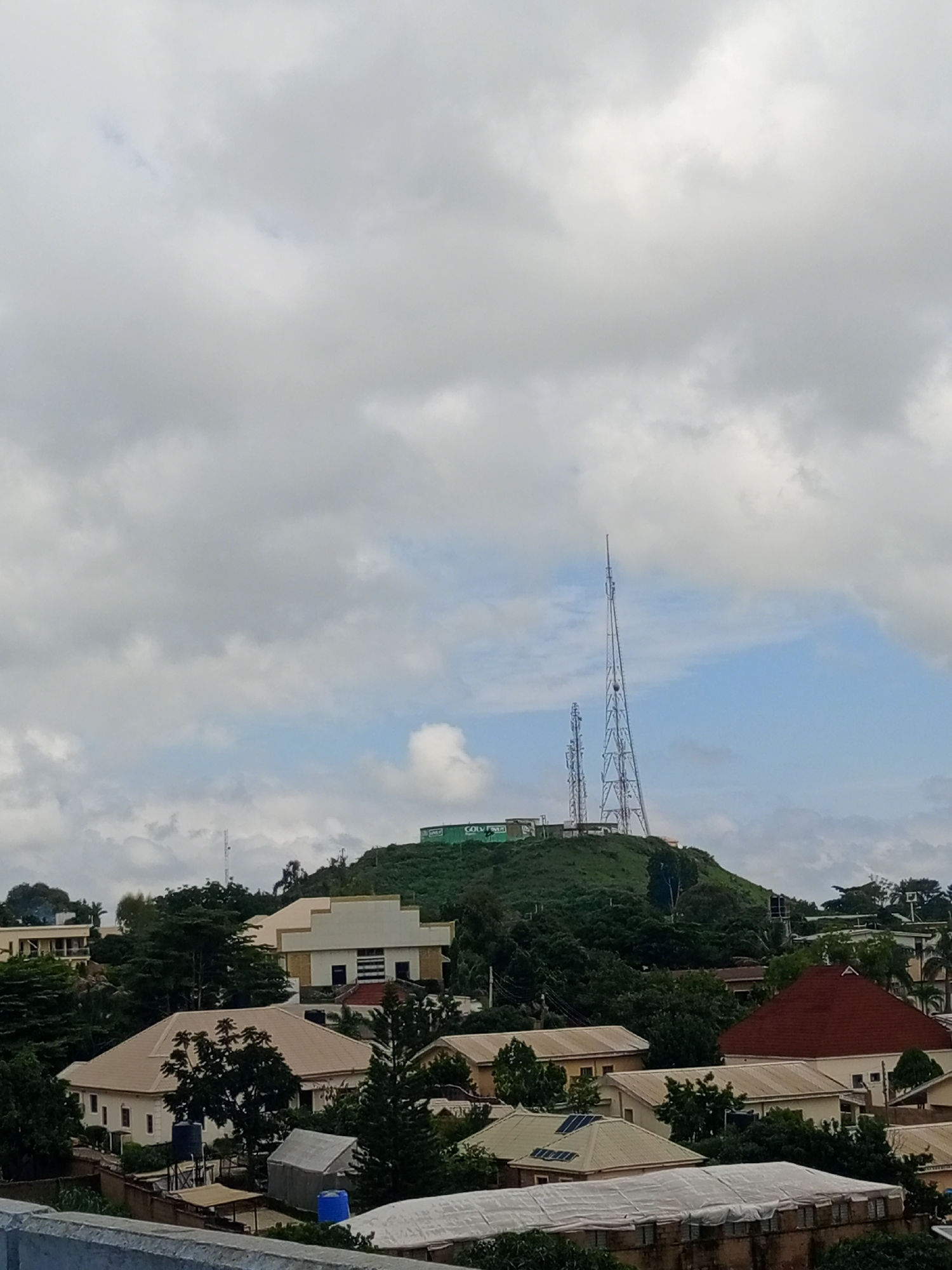 The image depicts a view of the city of Jos, Nigeria, looking towards a prominent hill. On top of the hill, there are two large communication towers, one significantly taller than the other, with a green sign visible on the summit. The hill is covered with lush green vegetation. In the foreground and mid-ground, there are several residential buildings with various roof types, including red and beige tiles. Some of these houses have solar panels installed on their roofs. Trees and other foliage are interspersed among the buildings, indicating a well-treed urban environment. The sky is partly cloudy with a mix of gray and white clouds, and patches of blue sky are visible. The overall impression is that of a daytime scene in a hilly urban area.