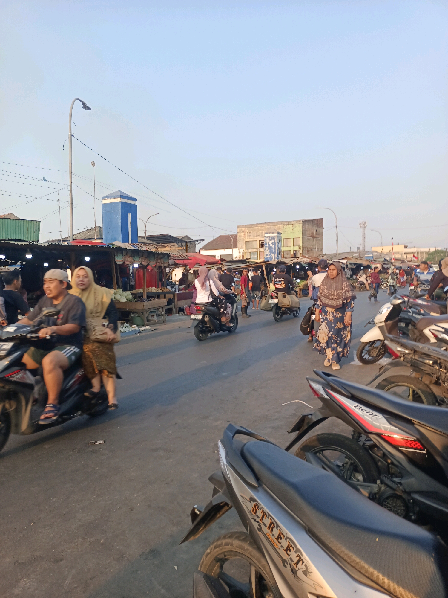 The image captures a bustling street scene, likely a marketplace, in Karawang, Indonesia. The setting is outdoors with a clear, pale blue sky overhead, suggesting daytime. A paved road forms the foreground and middle ground, occupied by numerous motorcycles, many parked, and some in motion. People are seen riding motorcycles, walking, and browsing stalls.
On the right side of the road, a row of market stalls with corrugated metal roofs is visible. These stalls display various goods, including produce like cabbages and other vegetables. Several individuals are engaged in transactions or moving through the market area.
In the background, various buildings of different sizes and colors, including a prominent blue and white structure with a pointed roof, can be observed. Utility poles and wires are also present, indicating an urban or semi-urban environment. The overall atmosphere is one of activity and commerce.
Specifically, the presence of motorcycles, the style of the market stalls, and the attire of the people, particularly the women wearing hijabs, are consistent with scenes found in Indonesia. The blue and white building might be a landmark or a government facility. The lighting suggests it is either early morning or late afternoon.
