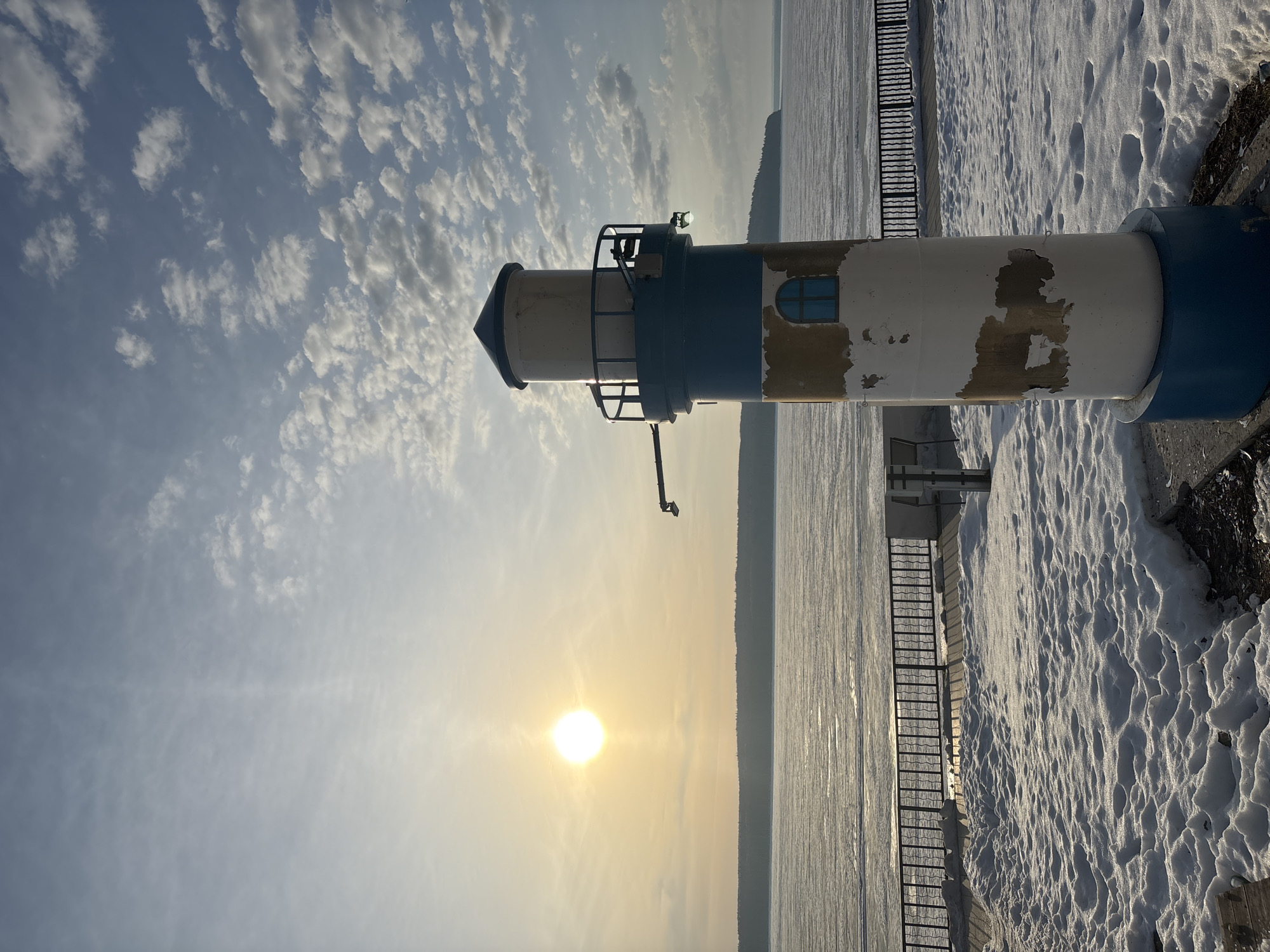 The image shows a daytime view of a lighthouse in the city of Chaikovsky, Russia, during winter. The lighthouse is painted with white and blue stripes and appears somewhat weathered. To the right of the lighthouse is a snow-covered area. In the background, a frozen body of water is visible, with trees along the far shore. The sun is shining brightly, creating a glare in the sky with patchy clouds above. A metal ladder is attached to the side of the lighthouse.