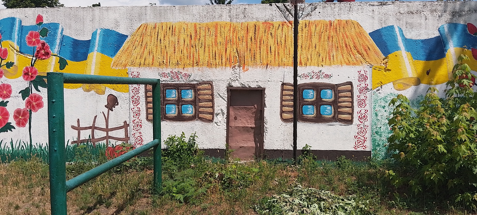 A mural on a white concrete wall depicts a traditional Ukrainian house with a thatched roof, two windows with blue panes and brown shutters, and a brown door. The house is flanked by stylized Ukrainian flags in blue and yellow. To the left of the house, red flowers with green stems and leaves are painted. To the right of the house, a green metal railing runs across the foreground. The mural is located in Cherkasy, Ukraine. The wall itself is textured and aged. In front of the wall, a patch of green grass and weeds grows, with a few small plants and bushes also present. The sky is overcast. The overall scene suggests a rural or historical representation within an urban setting.