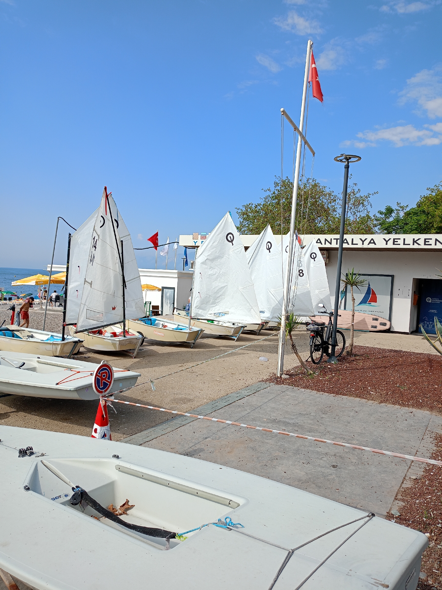 The image captures a sunny day scene at a beach in Antalya, Turkey, likely at a sailing club or school. In the foreground, several small, white sailboats are lined up on the sand and paved area. These boats appear to be for training or racing, with their sails either furled or partially raised. One boat in the foreground has a "P" parking symbol in a circle with a red border on a cone.
Further back, a building with the words "ANTALYA YELKEN" (Antalya Sailing) is visible, suggesting the location. The Turkish flag flies from a flagpole in front of the building. Several more sailboats are positioned behind the first row. In the background, beach umbrellas and beach chairs indicate a recreational beach area. People are present on the beach, some walking and others sitting. A bicycle is parked near the building. The sky is a clear blue with a few wispy clouds. The environment is a mix of sand, pavement, and some vegetation, characteristic of a coastal area.