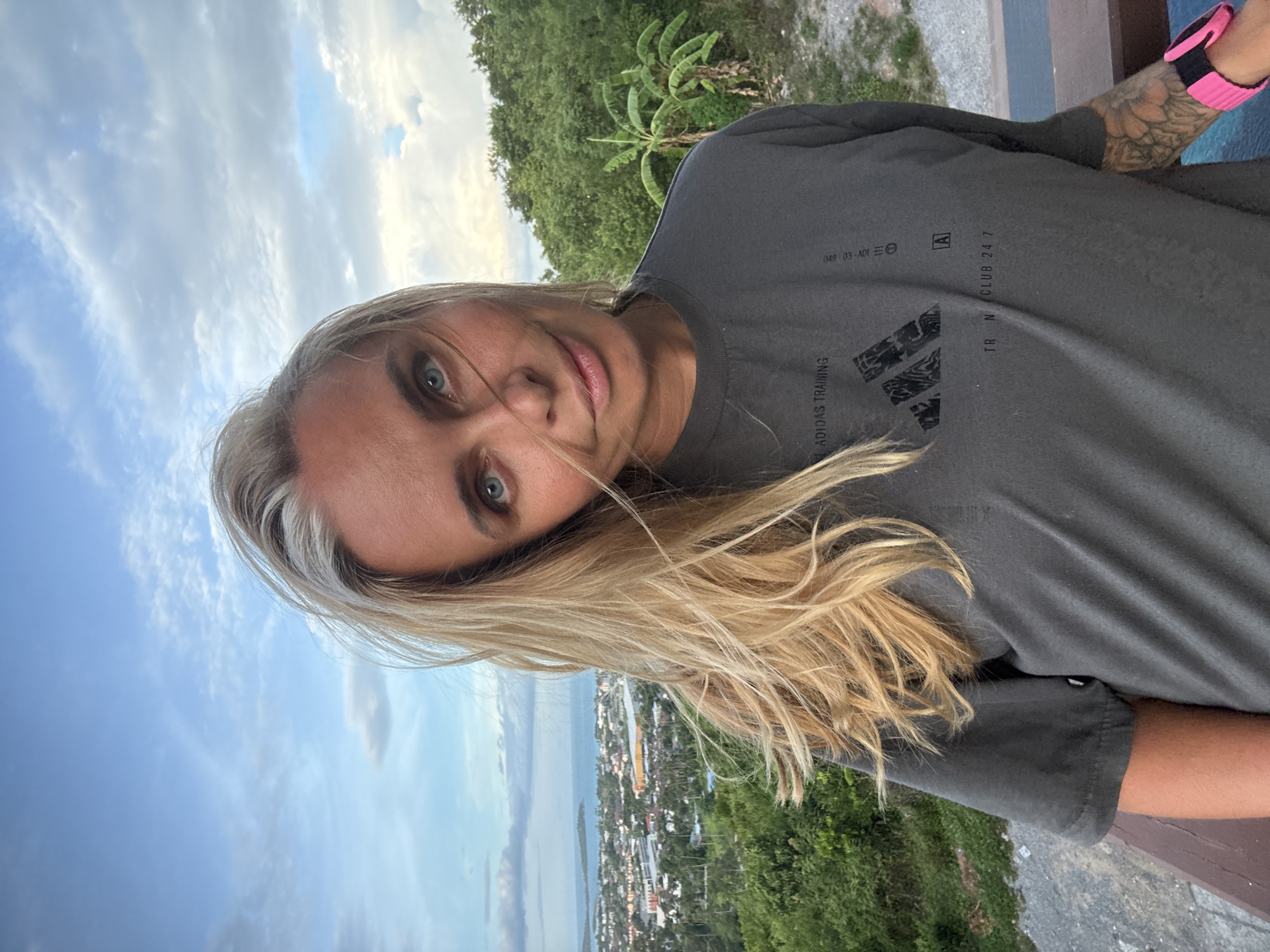 A young Caucasian woman with blonde hair and blue eyes is taking a selfie outdoors. She is wearing a dark gray Adidas t-shirt with "ADIDAS TRAINING" printed on the chest, along with the Adidas logo. Her right arm is visible, showing tattoos and a pink and black wristband.
The background shows a lush, green landscape with palm trees and dense foliage, typical of a tropical environment like Ko Samui, Thailand. In the distance, a town with buildings and houses can be seen bordering a body of water, likely the ocean, with a cloudy sky overhead. The lighting suggests it is either early morning or late afternoon, with soft, diffused sunlight. The woman's expression is relaxed and content. Visible text on her shirt includes "ADIDAS TRAINING" and "TRAIN. CLUB 24/7."