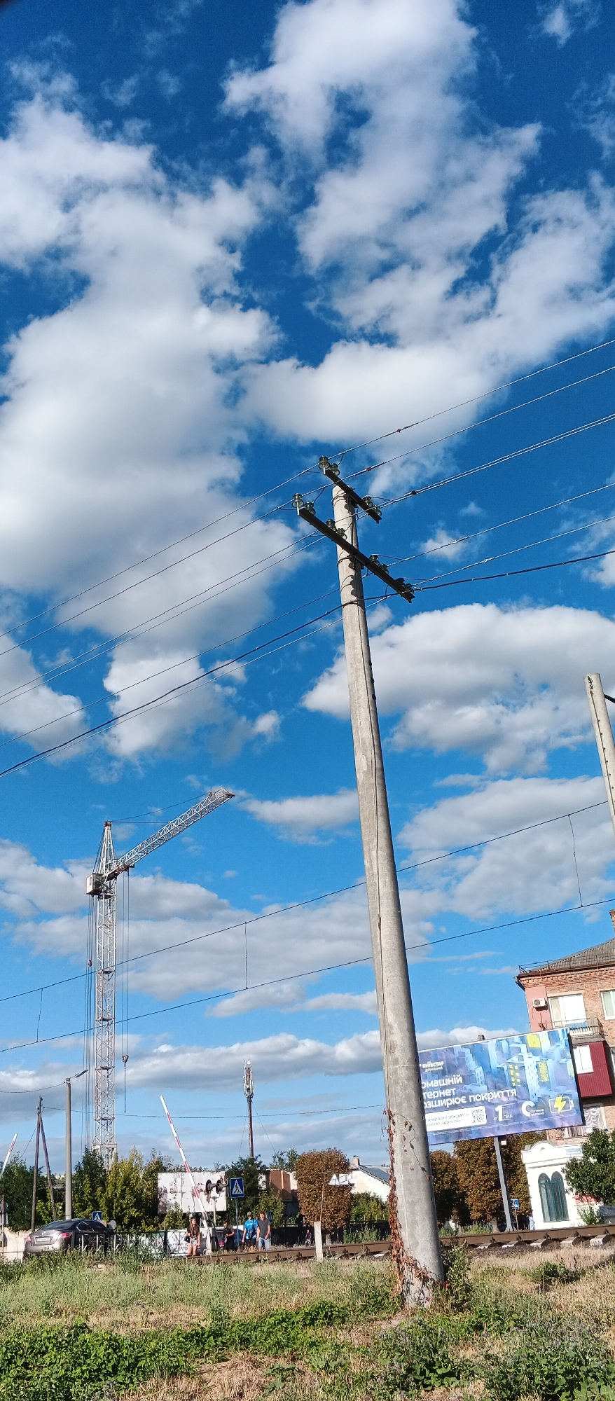 A daytime, low-angle, full shot captures a concrete utility pole in the foreground, leaning slightly to the right, with power lines extending horizontally across the frame. The background features a bright blue sky with fluffy white clouds. To the left, a construction crane is visible against the sky. In the middle ground, a railway crossing with white and red barriers is active, with several people standing nearby. A dark-colored car is parked on the left side of the crossing. Further back, trees and buildings are present. A billboard with Ukrainian text is mounted on the side of a red-brick building to the right. The scene appears to be in an outdoor, semi-urban environment, likely in the city of Smila, Ukraine, given the presence of construction and urban infrastructure.