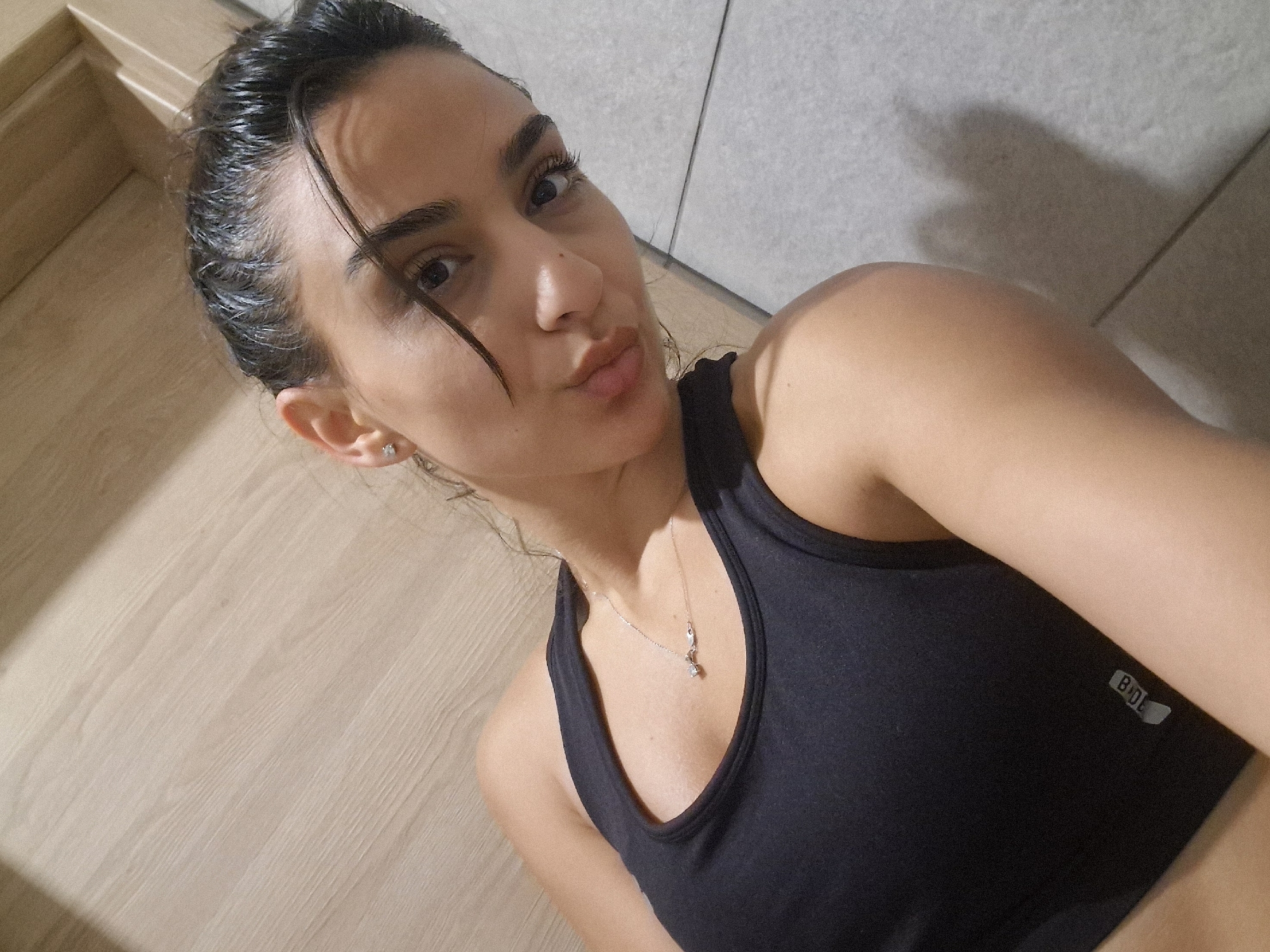 The image is a close-up selfie of a young woman in what appears to be a bathroom or changing room. She has dark hair pulled back and is wearing a black sports bra with a small logo on the side that reads "BODE". A delicate silver necklace with a charm is visible around her neck. Her expression is playful, with her lips pursed for a kiss. The background shows two distinct textures: a light-colored, wood-grain surface on the left and a gray, tiled surface on the right. There is a shadow cast on the tiled wall, possibly from the woman's arm or another object. The lighting suggests it is daytime, though the specific time is not discernible. There are no other people or objects of significant note in the frame.