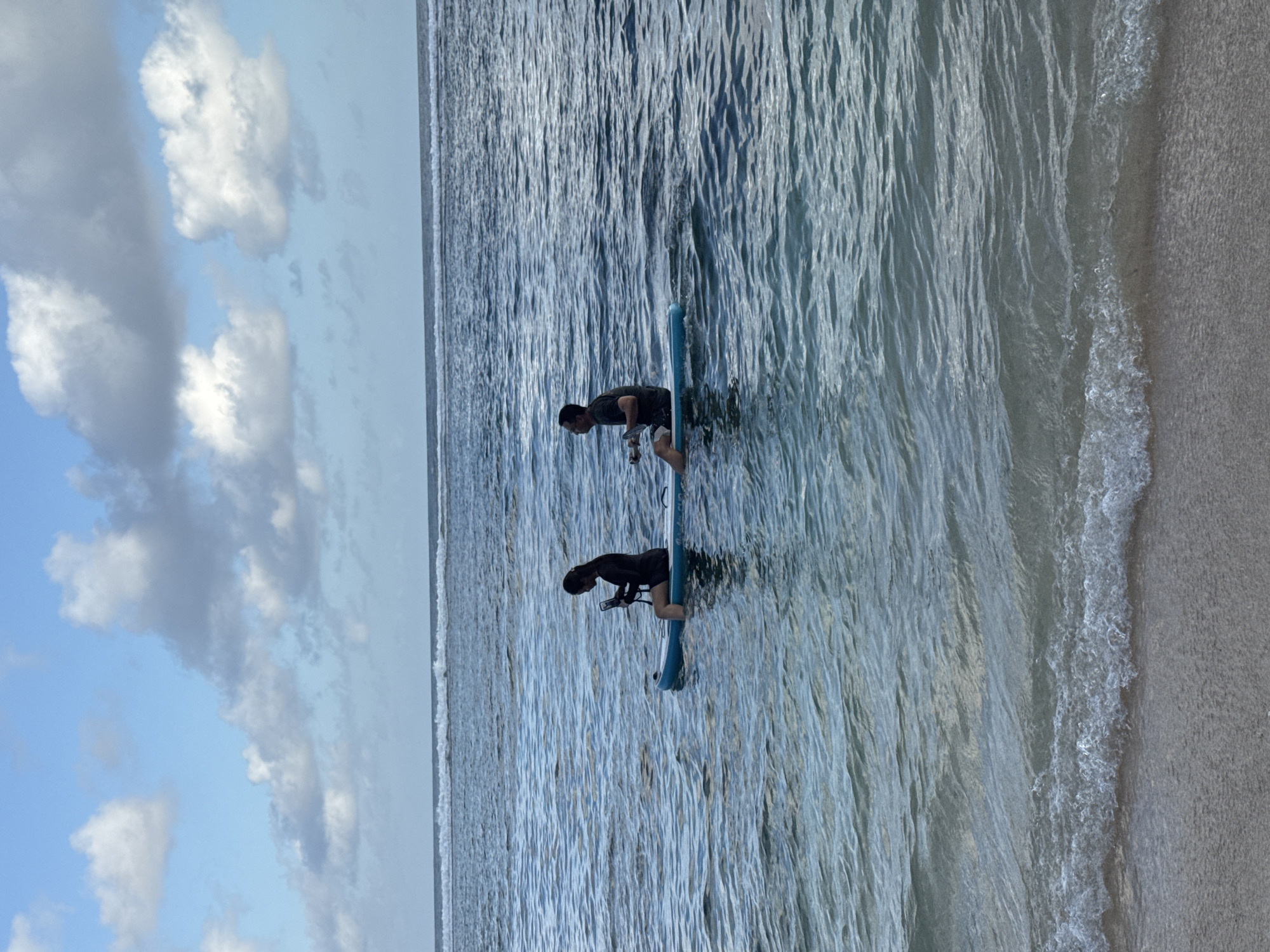 Two people are standing on a paddleboard in shallow water near a beach in Kuta Selatan, Indonesia. The water is clear and blue, and the sky is partly cloudy. One person is in the front of the paddleboard and is looking down at their phone or camera and the other is in the back. The beach is sandy and white.