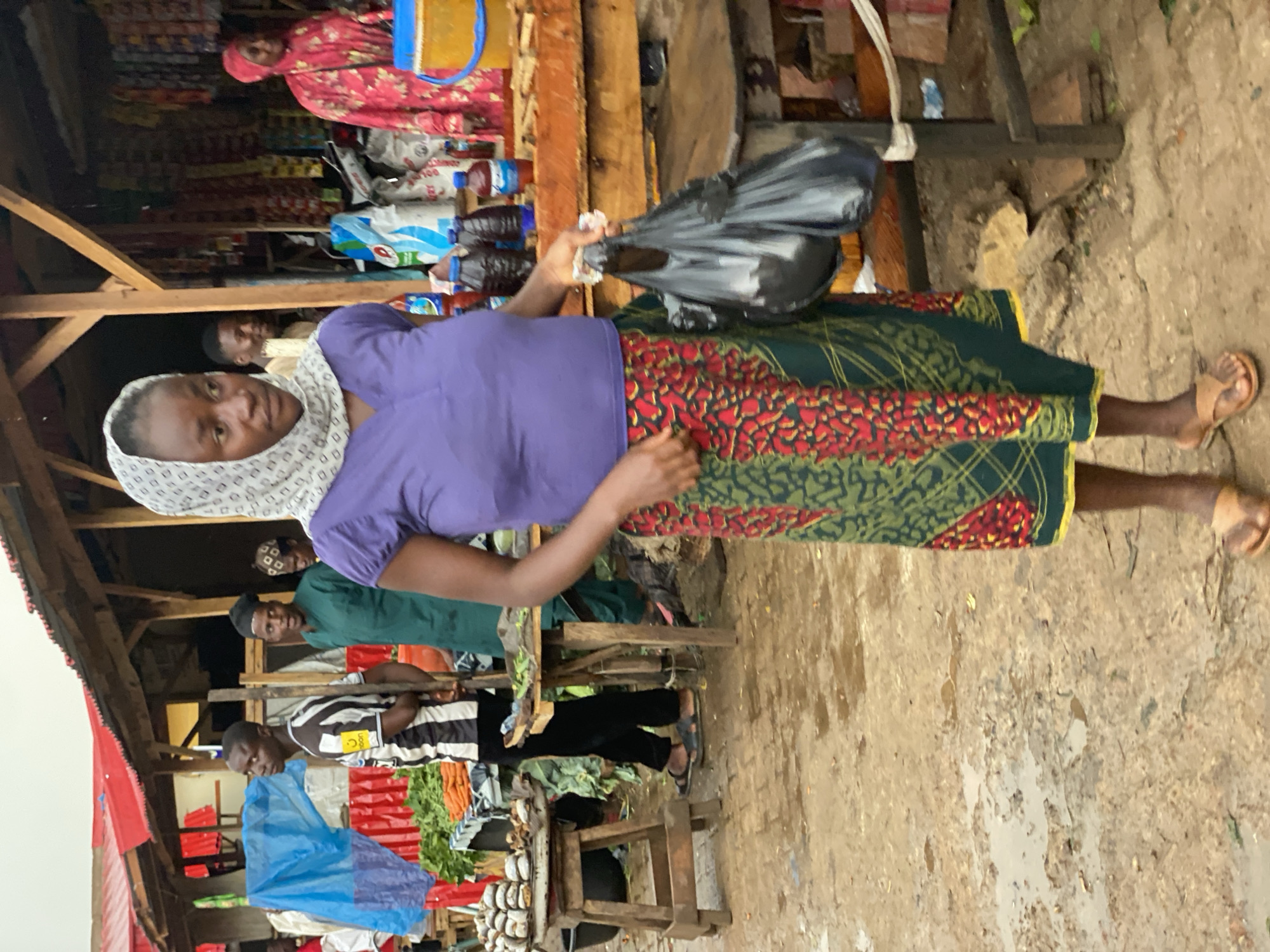 The image shows a marketplace in Kafanchan B, Nigeria. A woman in a purple shirt, patterned skirt, and headscarf is holding a black plastic bag. She is standing in a muddy area. Behind her, other people are present, including individuals near a wooden structure that appears to be a shop or stall. Vegetables and other goods are visible on display. The environment is generally weathered and appears to be an active commercial zone.