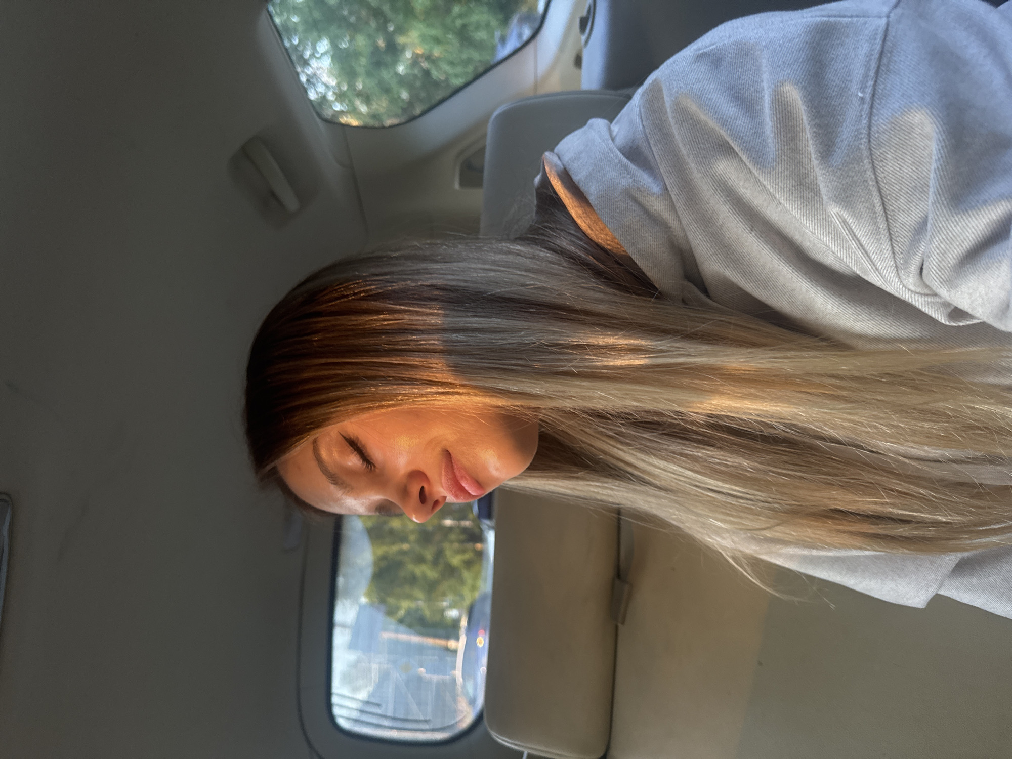 A young woman with long blonde hair is resting her eyes while sitting in the back seat of a car. Sunlight streams in from the side, illuminating her face and the side of her gray hoodie. She is looking downwards and to the left. The interior of the car is visible, with a headrest and a window showing trees and a road outside. The scene appears to be in the city of Staroe Mart'yanovo, Russia, possibly during a car ride in the late afternoon or early evening.