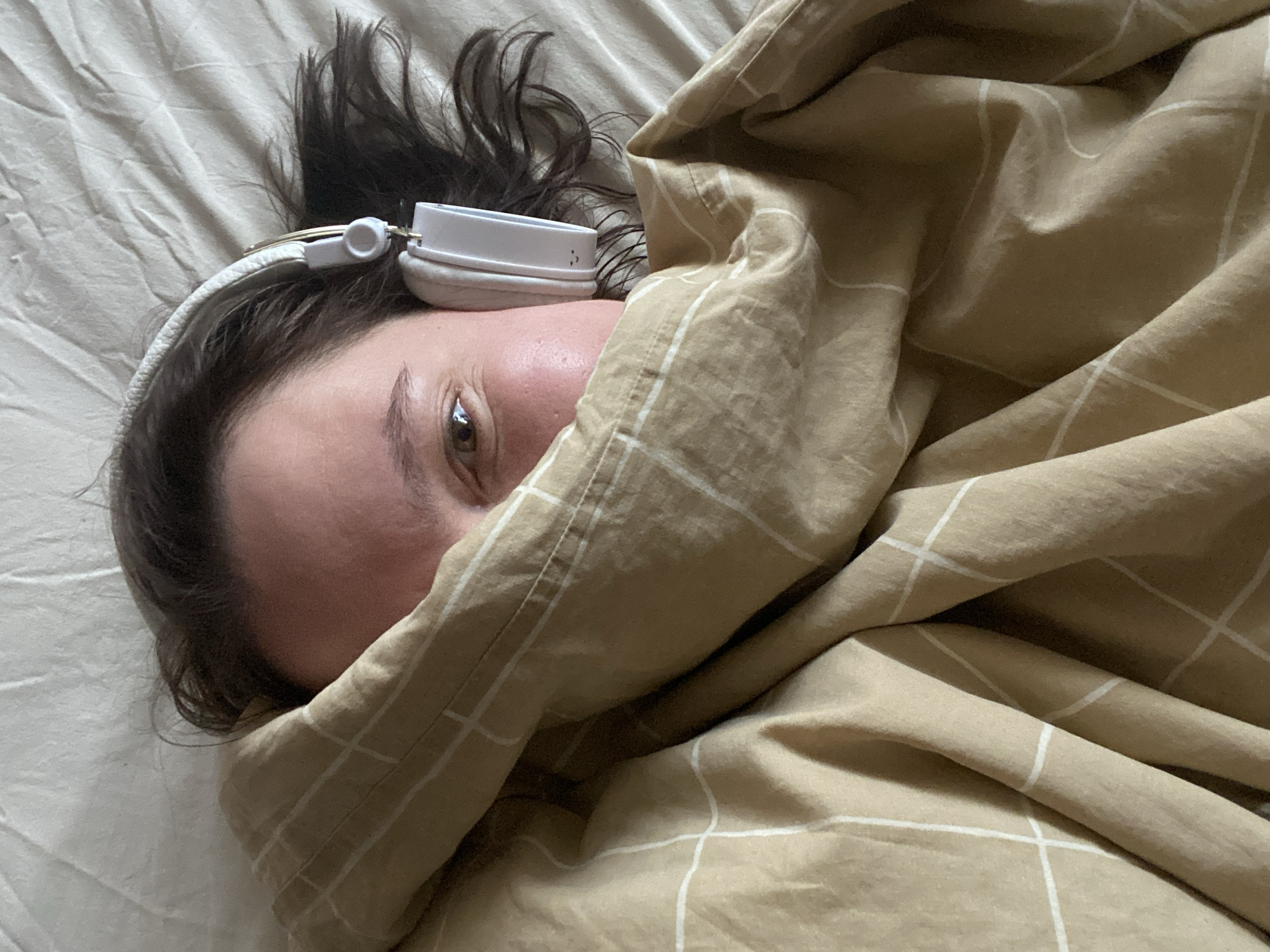 An individual is captured in a close-up, intimate shot, lying down indoors, likely in a bed. The person's face is partially obscured by a light brown or beige blanket featuring a subtle white grid pattern, creating a cozy and snug appearance. Only the left side of their face is visible, including an open brown eye that looks directly towards the camera, a portion of their forehead, and cheek. Dark brown hair, appearing somewhat disheveled, frames their head, extending onto a light-colored sheet or bed linen.
The person is wearing white or light-colored over-ear headphones, which rest comfortably on their head, suggesting they might be listening to music, a podcast, or an audiobook. On the visible earcup of the headphone, a small, dark, stylized symbol, resembling two slightly curved vertical lines or a minimalist audio icon, is discernible. The overall lighting is soft and natural, indicating an indoor environment, possibly a bedroom. The scene conveys a sense of relaxation or quiet leisure. This moment unfolds in Chaikovsky, Russia. No visible text beyond the headphone symbol is present.
