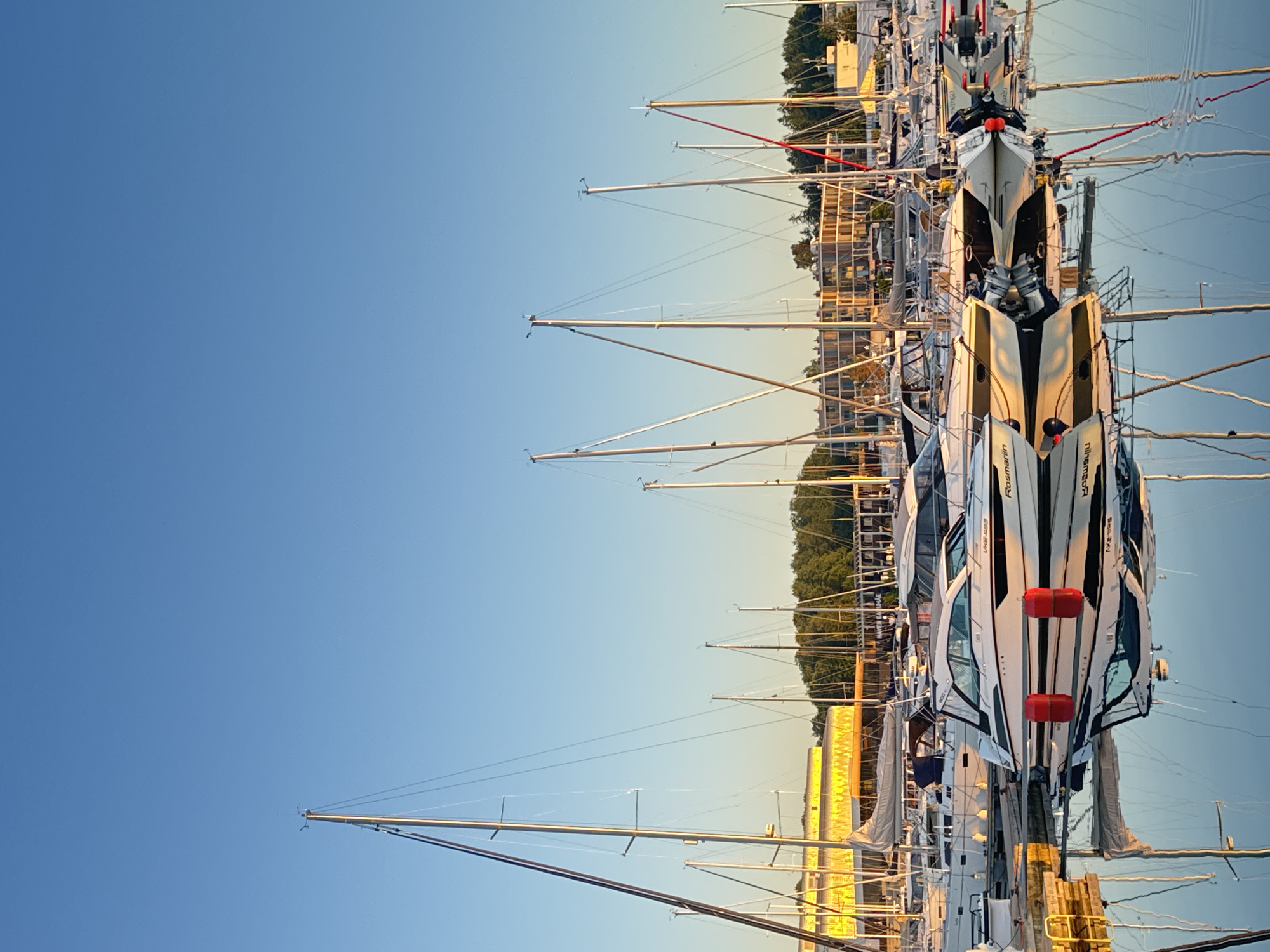 The image depicts a tranquil marina scene at Haven Kakumäe in Tallinn, Estonia, during either sunrise or sunset, characterized by warm, golden light. The upper portion of the frame is dominated by a clear, azure blue sky, transitioning to a lighter hue near the horizon.
Numerous boats are docked within the marina. In the foreground, a prominent white motor yacht, bearing the name "Rosmariin" and registration "VKG-493," is visible, along with another motor yacht displaying "R900." Both exhibit modern designs. Two red cylindrical buoys float in the water near the "Rosmariin" yacht. Surrounding these are various sailboats, identifiable by their tall, slender masts and rigging, one of which on the far right features a distinctive red section.
The water in the marina is exceptionally calm and still, creating nearly perfect, symmetrical reflections of all the boats, their masts, and the background structures. The reflections extend from the bottom third of the image upwards.
In the background, a distinctive multi-story building with a horizontally slatted, bright yellow/golden facade is prominent, identifiable as part of the Haven Kakumäe complex. To the right of this building, a cluster of green trees and other less distinct structures are visible in the distance. The warm, low-angle sunlight casts long shadows and highlights the illuminated sides of the boats and buildings. No people or active human presence are discernible within the frame; the scene conveys a sense of quiet stillness.