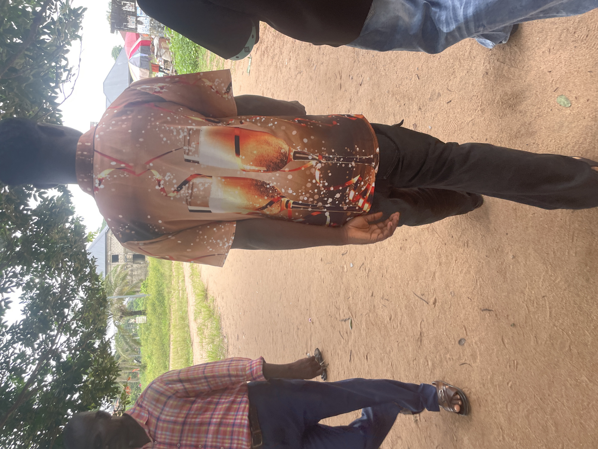 The image depicts three individuals walking on a dirt path in Ihiagwa, Nigeria. The person in the foreground, seen from behind, wears a short-sleeved shirt with a graphic print of two champagne glasses, bubbles, and streamers. This individual is also wearing dark pants. To the left, another person is walking away from the camera, wearing a plaid long-sleeved shirt in shades of red and pink, and navy blue pants. This person is wearing brown sandals. The third individual, partially visible on the right, is wearing a dark shirt and blue jeans. The environment is outdoors, with a dirt path and sparse vegetation. Trees with dense foliage provide shade overhead. In the background, there are buildings with visible construction and scattered umbrellas, suggesting a marketplace or community area.