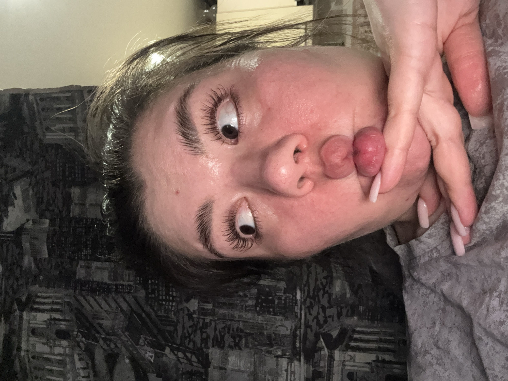 A person with light skin is positioned in the frame, with dark hair pulled back from their face and dark eyebrows. Their eyes are wide and looking upward, and they are puckering their lips in a pout. A hand is positioned to the side of the face, and the background appears to be a dark-patterned surface on the left and a light gray patterned surface on the right. There is no indication of the city of Votkinsk, Russia, or any geographical location.