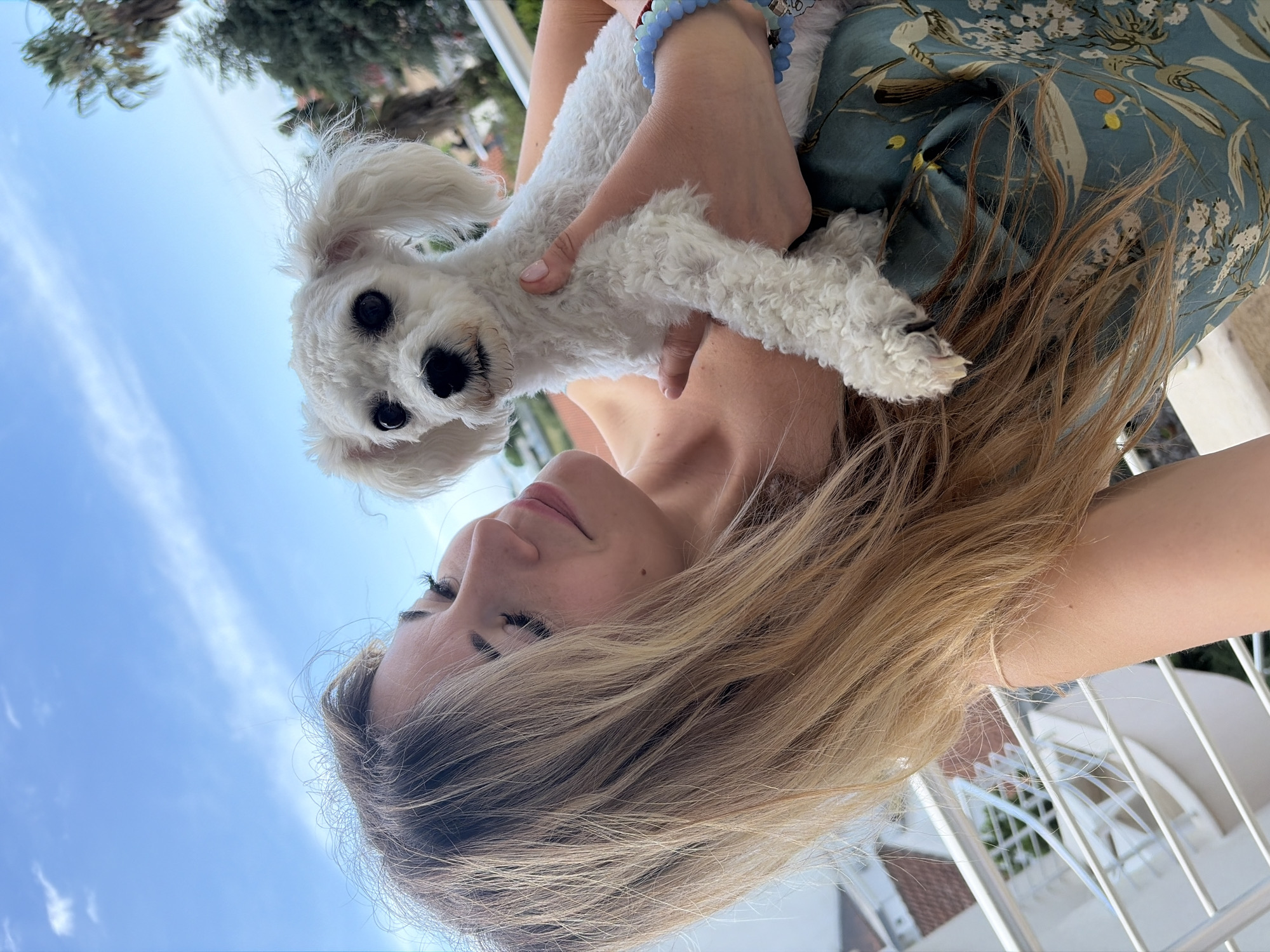 A woman is holding a small, white, fluffy dog. The background consists of a clear, blue sky and vegetation. The woman has long, blonde hair and is wearing a blue, floral-patterned top. She is also wearing a blue beaded bracelet on her wrist. The dog has black eyes and a black nose. The setting appears to be outdoors, with a white, wrought-iron railing visible, suggesting a balcony or similar structure. Considering the presence of a railing and the style of architecture in the background, the location is consistent with a residential area in Budva, Montenegro.