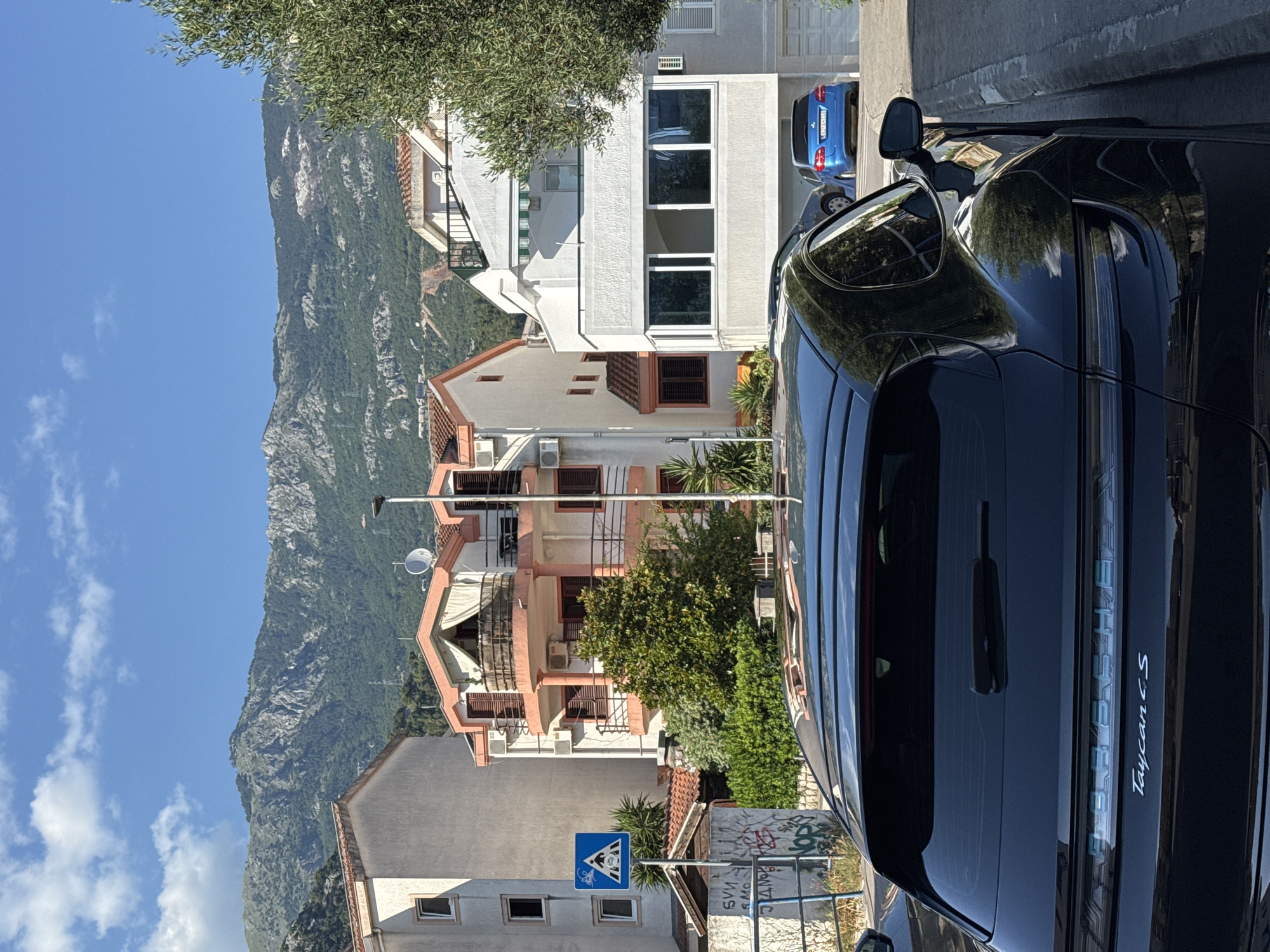 A black Porsche Taycan S is parked on the right side of a paved street in Budva, Montenegro, facing away from the camera. The car's rear windshield, spoiler, taillights, and "Porsche" and "Taycan S" badging are visible. The car is positioned next to a curb and a sidewalk. In the background, several multi-story buildings with peach and white facades are visible. A tall, green tree stands to the right of the frame. Beyond the buildings, a mountain range covered in green vegetation and rocky outcrops dominates the horizon under a clear blue sky with scattered white clouds. A blue hatchback is parked in a driveway on the right side of the street, further down from the Porsche. A blue pedestrian crossing sign with figures and a bicycle is posted on the left side of the street, in front of a graffiti-covered wall. The overall scene depicts a sunny day in a residential area with a view of mountains.