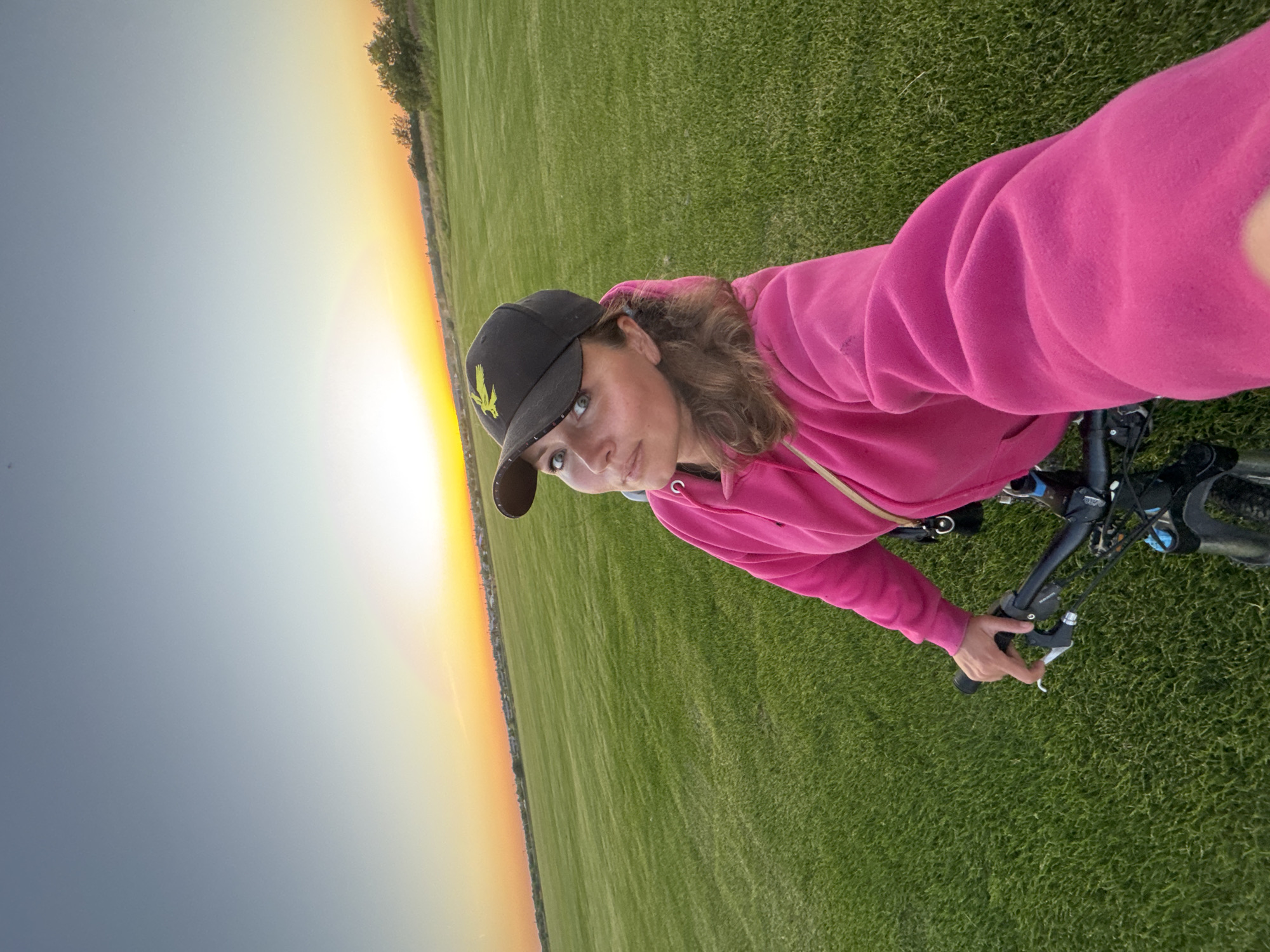 A woman is pictured from the chest up, riding a bicycle on a vast, well-maintained green field. She is wearing a bright pink hoodie and a black baseball cap with a yellow emblem. The sky above is a gradient of blue, transitioning to a warm orange and yellow hue near the horizon, indicating either sunrise or sunset. In the distance, a line of trees and some buildings can be seen. The image has a slightly wide-angle perspective, capturing the expansive nature of the field and the sky. The camera appears to be held by the subject herself, creating a selfie-style composition. The location is identified as Kempelevo, Russia.