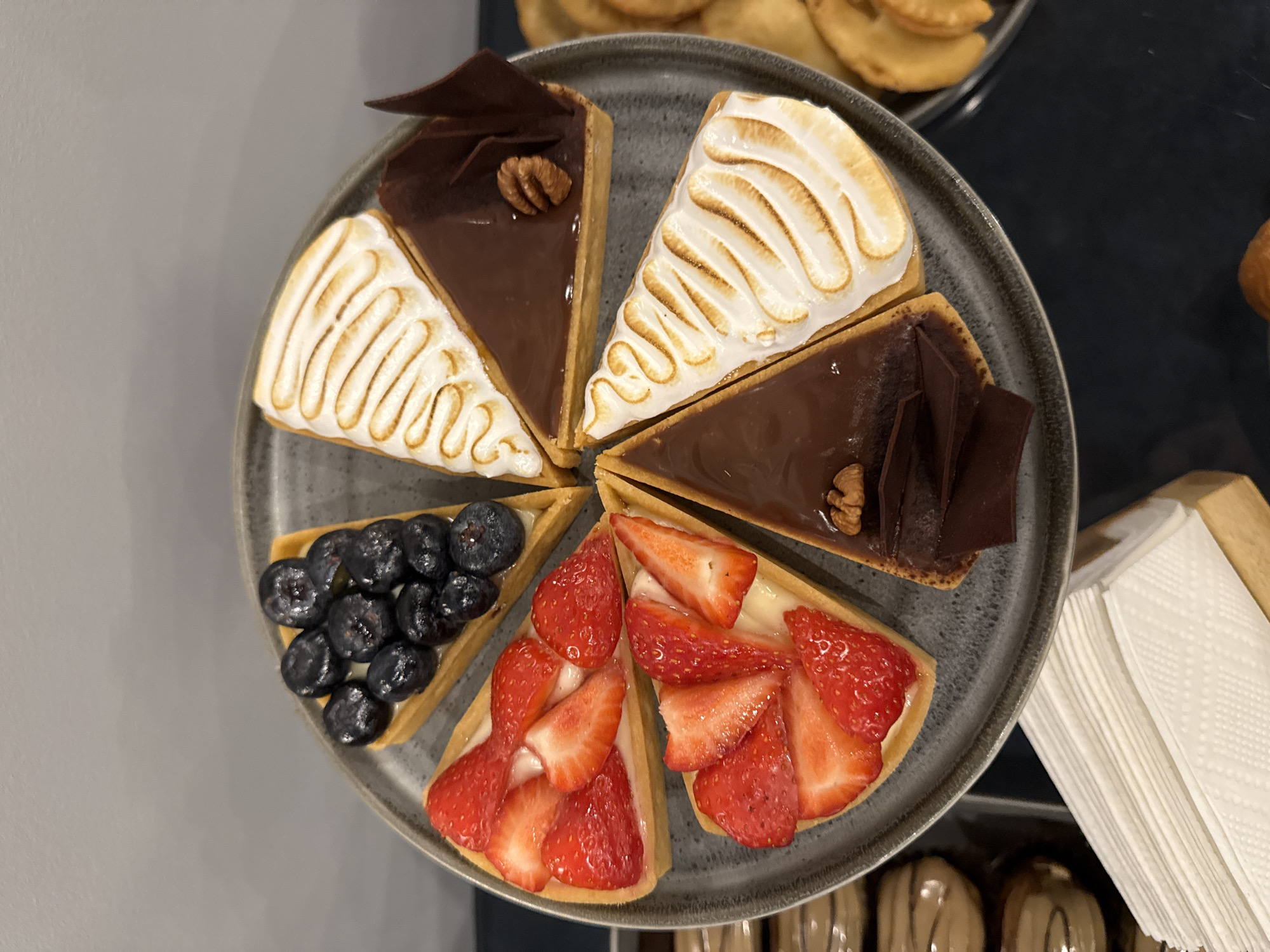 The image shows a gray plate with six triangular pieces of assorted tarts. Two pieces have chocolate filling, chocolate shavings, and a walnut. Two pieces are topped with torched meringue. One piece is covered in blueberries and another in sliced strawberries. A stack of white napkins sits next to the plate. In the background, there is a plate with small pastries and another sweet treat with a glazed topping. The image was taken in Perm, Russia.