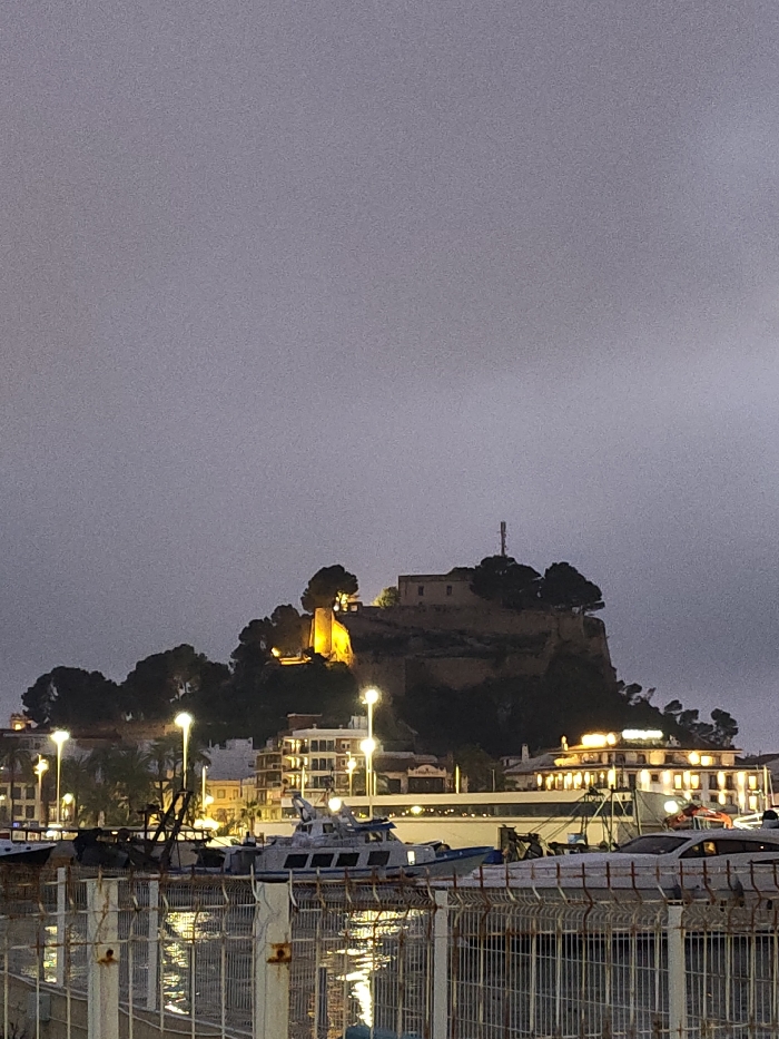 The image depicts a nighttime or dusk scene in Dénia, Spain, overlooking the city's port and the Dénia Castle. The sky is dark grey and overcast, occupying the upper two-thirds of the frame.
In the midground, the Dénia Castle is prominently situated atop a steep, rocky, and heavily treed hill. Portions of the castle structure are illuminated with warm yellow light, highlighting a tower on the left side of the castle. A tall, slender communication tower is visible on the highest point of the castle grounds. The hillside below the castle is dark, with silhouetted trees.
Below the castle, a cluster of multi-story buildings lines the waterfront. Many of these buildings have illuminated windows, emitting warm light. One building on the right features illuminated signage, possibly reading "HOTEL." Numerous streetlights with bright yellow-white illumination are spaced along the waterfront, casting reflections on the water.
In the foreground, a body of water is visible, likely the port, with reflections of the city lights. Several boats are docked or moored in the water; one prominent white and blue vessel with multiple windows is visible slightly left of center. The immediate foreground is dominated by a metal fence, possibly a security or construction barrier, with vertical posts and horizontal rails. No people or specific actions are discernible within the frame.