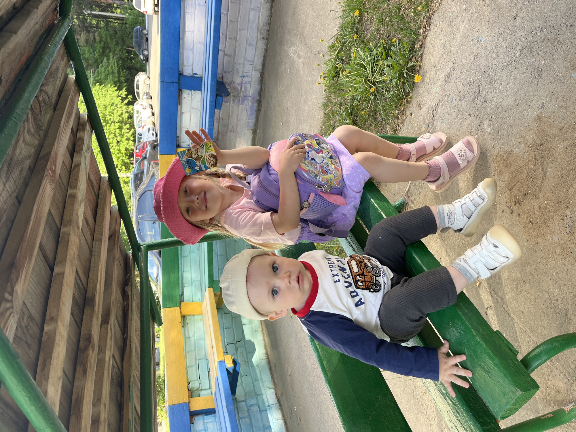 Two children are sitting on a green bench at an outdoor playground. A girl with blonde hair, wearing a pink hat, pink shirt, and purple dress, has a purple backpack behind her and is holding a small card. A younger boy with blue eyes and a light-colored hat is wearing a long-sleeved shirt with a car design on it and dark leggings. The background features a colorful play structure with yellow and blue painted surfaces, a concrete walkway, and some greenery. The location is most likely a playground in the city of Chaikovsky, Russia.