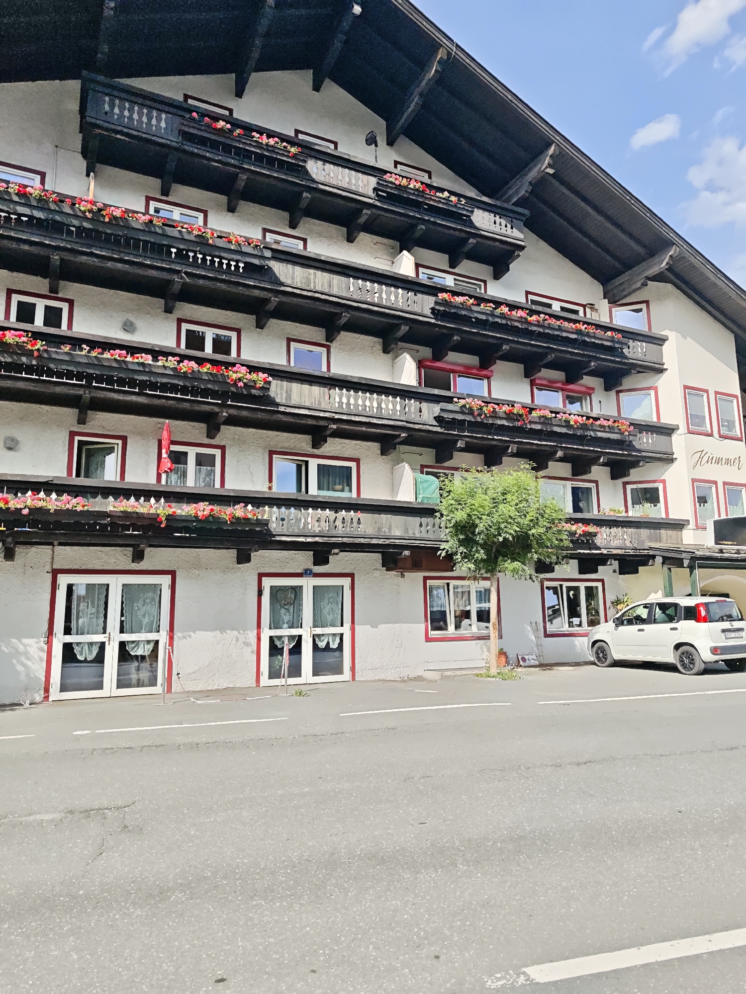 The image shows the exterior of a multi-story building with a dark roof and balconies adorned with red and pink flowers. The building is painted a light color and has red trim around its windows and doors. A white car is parked on the street in front of the building. A tree stands to the left of the car. The building has signage that reads "Hümmer." This is a scene from Kitzbühel, Austria. The sky is blue with scattered clouds. The street is paved and has a white line marking a lane.