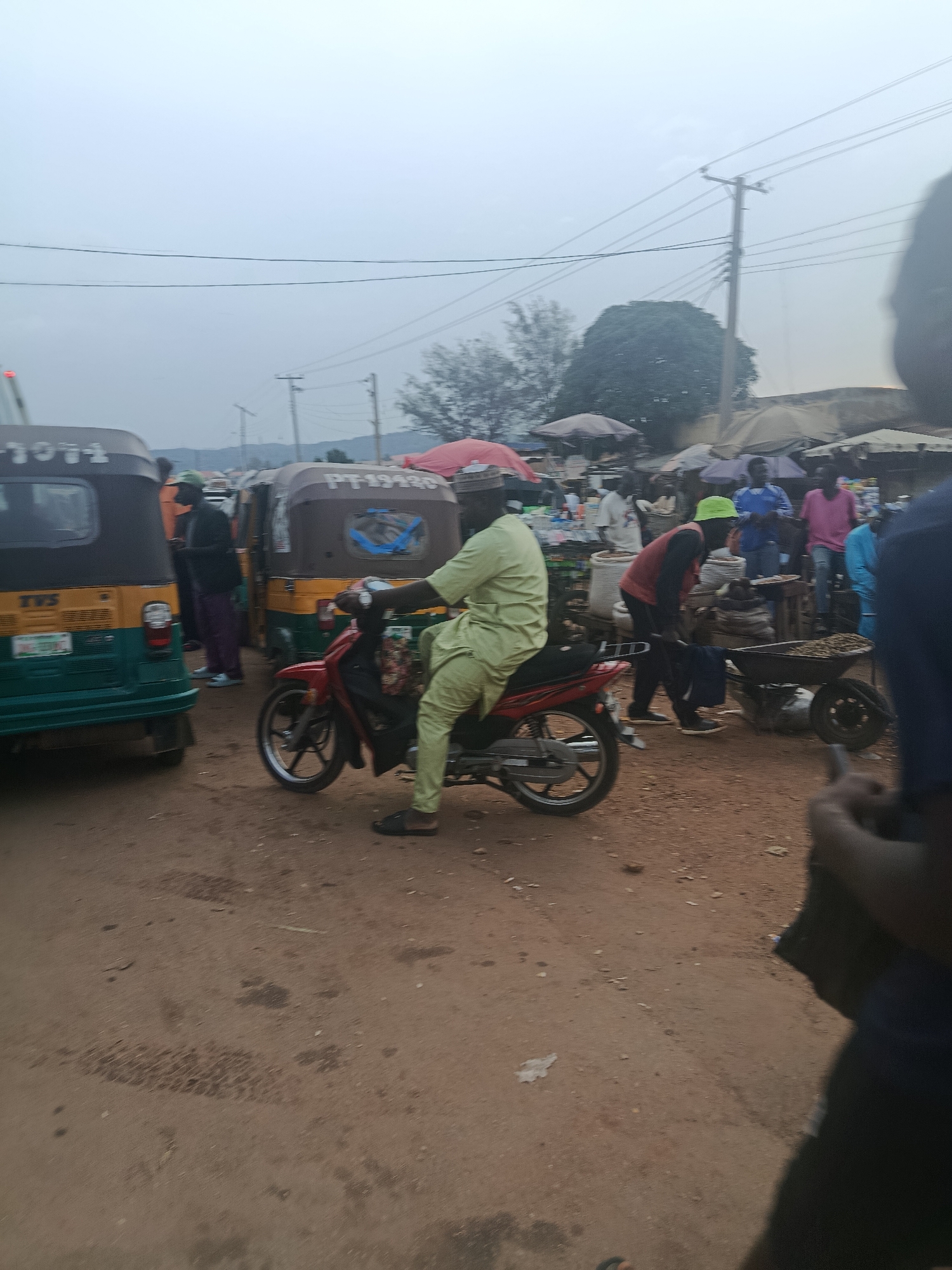 The image depicts a bustling outdoor market scene in Rafin Par, Nigeria. The foreground is dominated by a red motorcycle with a rider wearing a light green traditional outfit. The motorcycle is on a dirt road. Behind the motorcycle, two auto-rickshaws are visible. The one on the left is green and yellow, with "TVS" visible on its side. The one on the right is brown and yellow, with "PT-19920" written on it.
Several people are present in the background, some appearing to be vendors and customers. Some individuals are wearing brightly colored clothing, while others are in more subdued tones. Tents and market stalls are set up, suggesting a marketplace environment. In the distance, trees and a cloudy sky are visible. Power lines are strung across the upper portion of the image. The ground is a reddish-brown dirt. One person in the mid-ground, wearing a red vest and a light green cap, is pushing a wheelbarrow filled with what appears to be produce or goods. The overall impression is one of a vibrant, active community market.