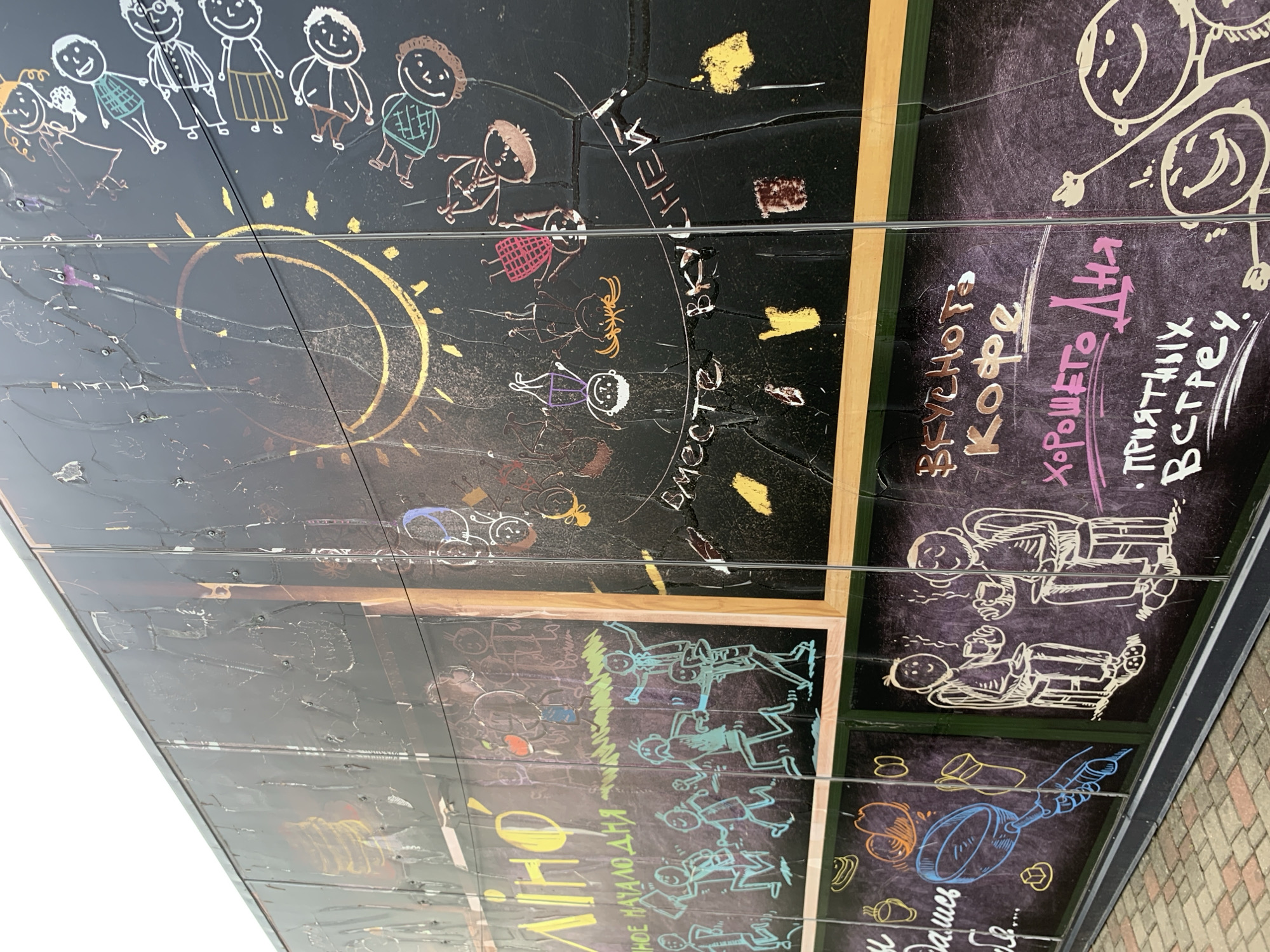 The image shows a glass exterior wall of a building, likely a cafe or shop, with chalk-like drawings on it. The drawings depict stick figures of children in various poses, some appearing to be dancing or celebrating. There are also drawings of a sun or moon, food items like apples and coffee cups, and text in Cyrillic. The overall style is playful and childlike. The location is identified as Sluck, Belarus, suggesting this is a local establishment. The glass appears to be segmented, with visible seams between panels, and there are some reflections on the surface. The drawings are rendered in a variety of colors, including yellow, blue, pink, and white.
