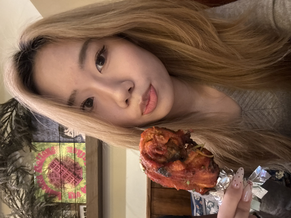 A young adult female, appearing East Asian, is depicted from the upper torso, holding a large, cooked, red-orange chicken drumstick wrapped in aluminum foil in her left hand. Her light blonde, layered hair, with visible dark roots, frames her face. She has dark eyes with visible eyeliner and light pink, slightly pursed lips, looking directly at the camera. She wears a light gray V-neck ribbed sweater. Her left hand displays almond-shaped fingernails with a light polish featuring red dots and a cherry design. The background indicates an indoor dining establishment located in Yuzhong District, China. A wooden shelf behind her head displays a square artwork with a concentric green, pink, and yellow waffle-like pattern. To its right are partially visible record albums or books, one mentioning "RAY CHARLES" and another featuring a partial face. Dried dark foliage is positioned along the top edge of the shelf. Below the shelf, a light-colored wall and dark wooden paneling are visible, alongside a blue and white patterned fabric on the lower left. The lighting is warm.