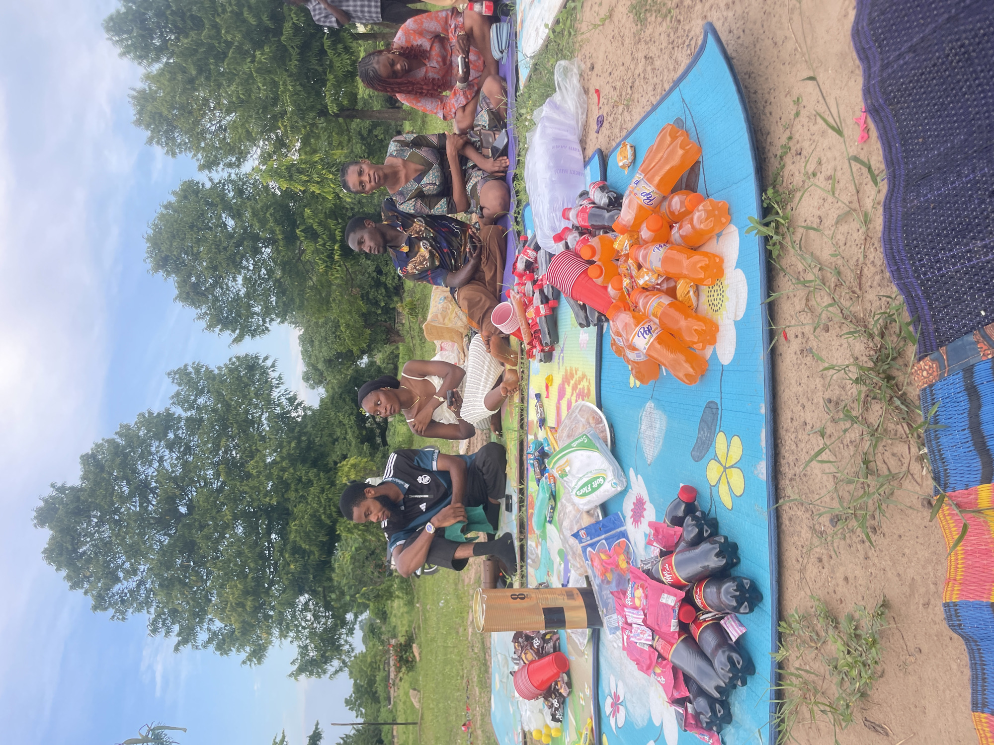 A group of six individuals, five seated and one standing, are gathered for a picnic on a grassy area with scattered trees in the background. The seated individuals are on blue and patterned mats laid out on the ground. In the foreground, a spread of snacks and drinks includes numerous bottles of orange soda and dark soda, individually wrapped candies, and bags of chips. A large cylindrical object, possibly a container or a cooler, stands upright among the items. One man, wearing a black and white soccer jersey, is seated with his legs crossed, looking to his left. Next to him, a woman in a white dress is looking at her phone. To their right, another woman in a patterned green and black dress is seated, and beside her, a woman in an orange and black patterned dress is also looking at her phone. The environment appears to be a park or an outdoor recreational area, likely in Lafia, Nigeria, given the vegetation and general landscape. The sky is partly cloudy, suggesting an outdoor event during the day. The standing individual is partially visible on the far right of the frame.