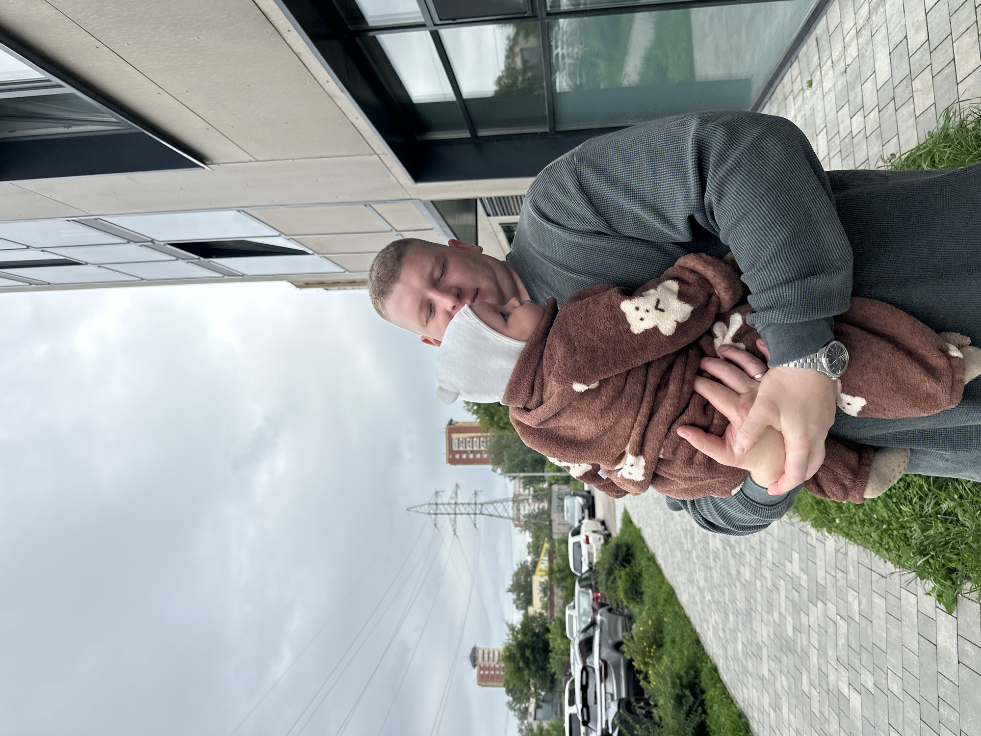 In Perm, Russia, a man in a grey long-sleeved shirt holds a baby wearing a brown fuzzy onesie with white teddy bear patches and a white bonnet. The man is looking down at the baby. They are standing on a paved walkway next to a grassy area. To their right is a modern building with a beige facade and large windows. In the background, there is a parking lot with several cars, trees, and power lines stretching across the cloudy sky. Two red brick buildings are visible in the distance, along with a tall power pylon. The scene appears to be a residential or commercial area in the city.
