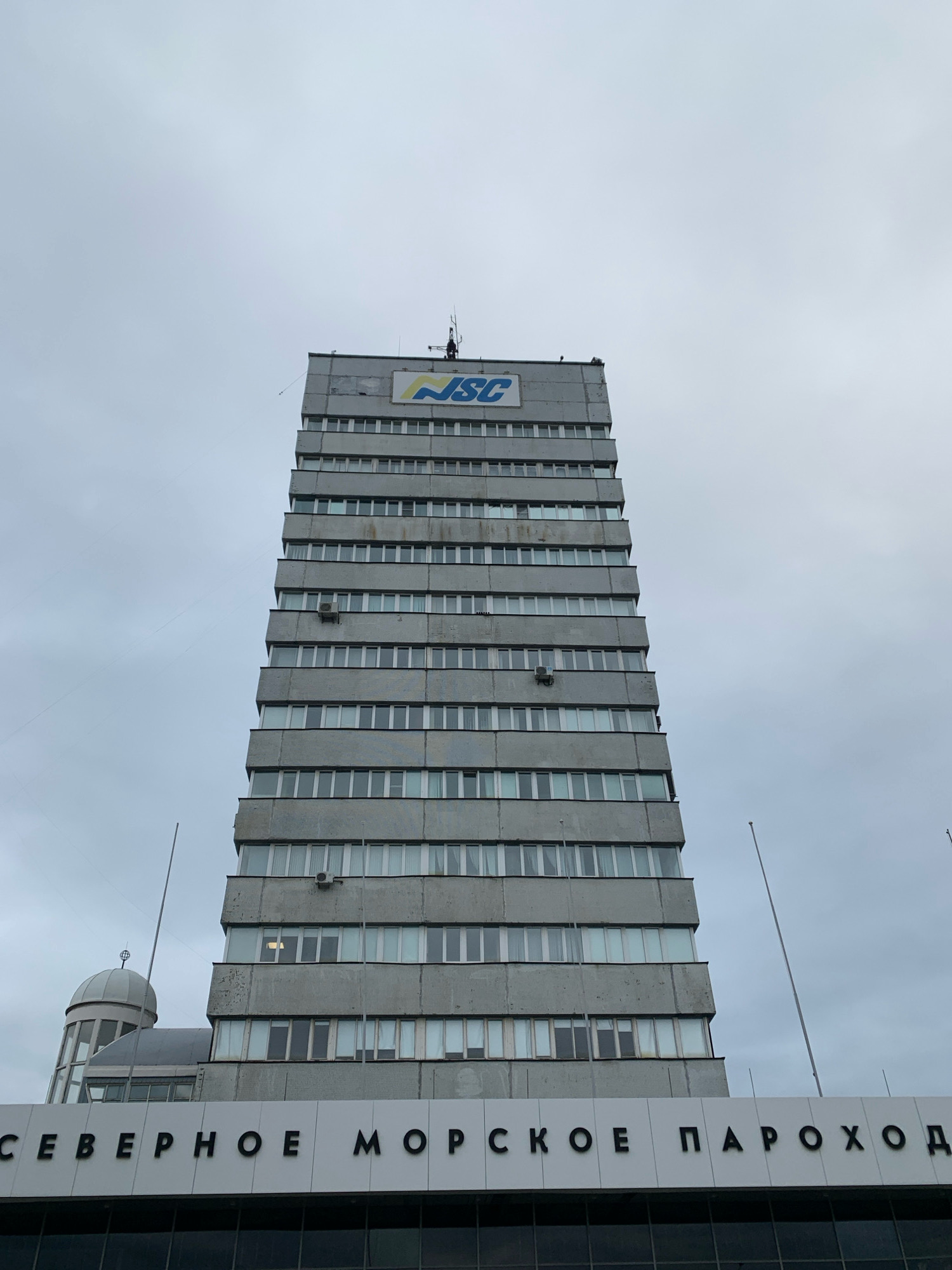 The image shows a tall, gray, Brutalist-style office building, likely in Arkhangelsk, Russia. The building features a repeating pattern of windows and concrete horizontal and vertical lines. At the top of the building, a blue and yellow logo with the letters "NSC" is visible. The bottom of the building is partially obscured by a white facade with large black Cyrillic lettering that reads "СЕВЕРНОЕ МОРСКОЕ ПАРОХОД". This translates to "Northern Sea Shipping". To the left of the main building, a smaller, domed structure is visible. The sky is overcast and gray. There are no people visible in the image. The scene appears to be a street-level view of the building's exterior on an ordinary day.