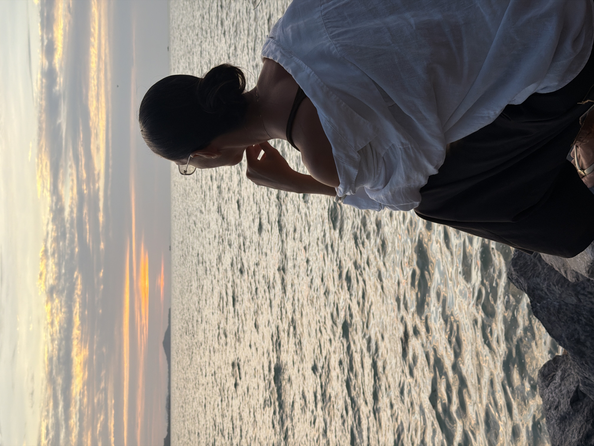 A lone woman with her hair in a bun, wearing a white shirt and dark pants, sits on rocks by the sea in Ko Samui District, Thailand. She is looking out at the ocean as the sun sets, casting a warm glow on the water and the cloudy sky. Her hand is resting on her chin, suggesting a contemplative pose. The waves gently lap against the shore, and the distant horizon is marked by a faint outline of land. The overall scene evokes a sense of tranquility and reflection.
