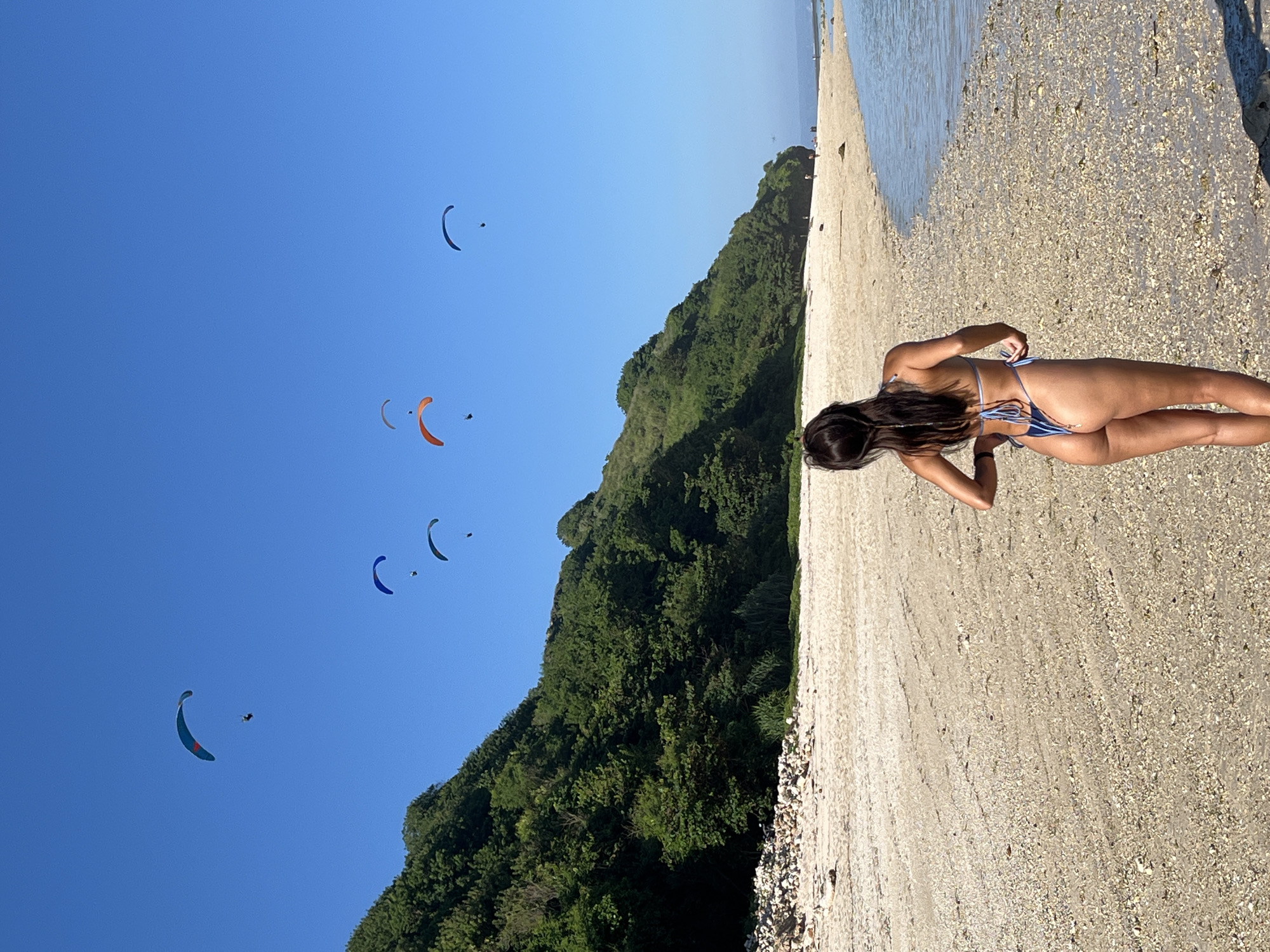 The image, displayed with a 90-degree clockwise rotation, captures a vibrant coastal landscape in Kuta Selatan, Indonesia. When viewed upright, the scene features a bright, clear blue sky dominating the upper left, transitioning to a lush, forested hill along the center, and a wide sandy and pebbly beach on the right, bordering the ocean.
In the foreground on the beach, a woman is seen from behind, facing away from the viewer. She has long, dark hair and is wearing a blue and white striped bikini, appearing to be standing or bending slightly, possibly adjusting her attire or looking out towards the horizon.
In the vast, clear blue sky, multiple colorful paragliders are visible, some with small figures below them, indicating active paragliding. The water of the ocean or sea is visible along the far right edge of the beach, creating a serene backdrop. The bright sunlight and absence of clouds suggest a clear, sunny day, likely during midday or early afternoon, with excellent visibility. The setting conveys a sense of outdoor leisure and adventure. There is no visible text in the image.