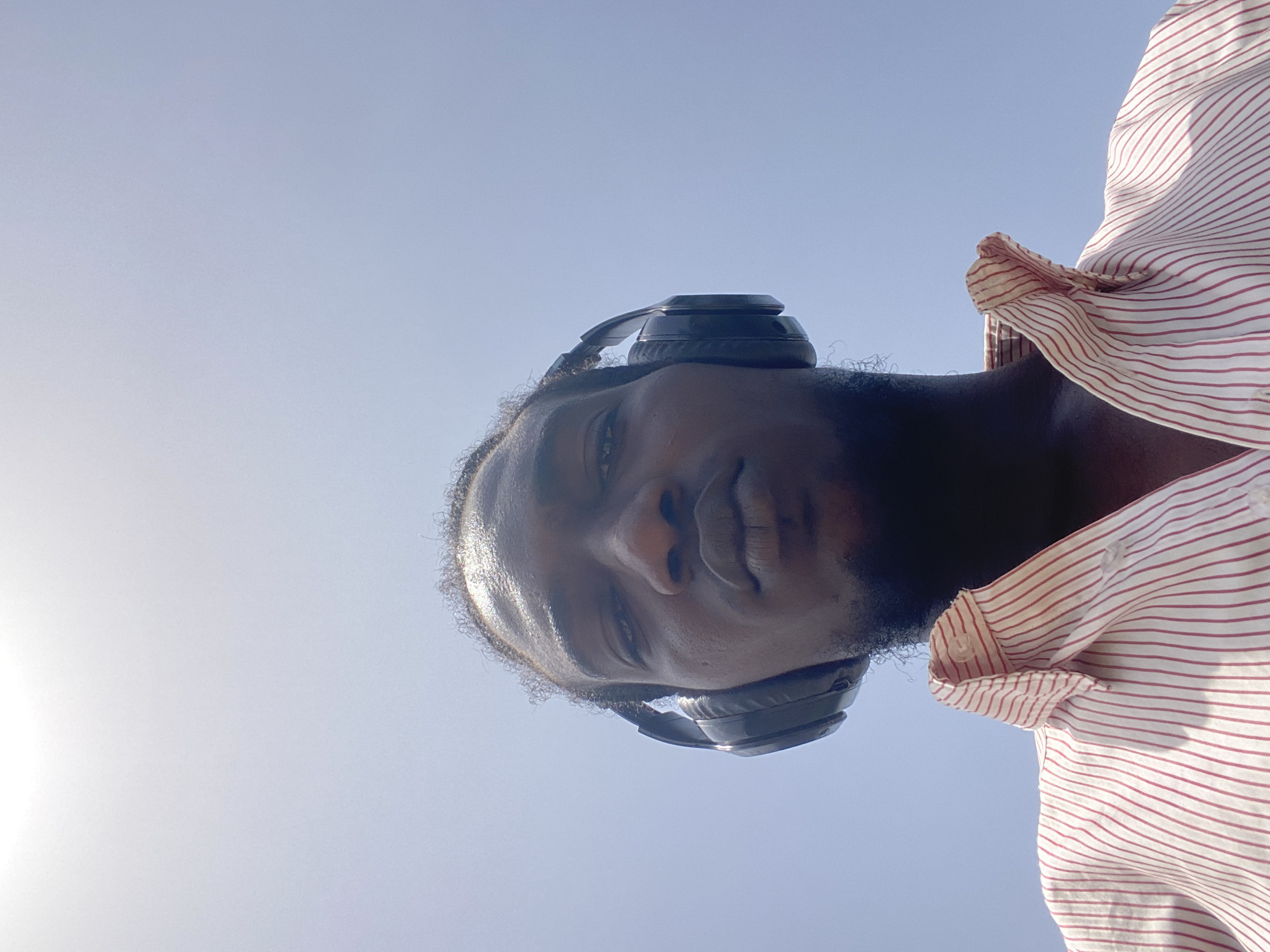 A low-angle shot shows a dark-skinned man with headphones against a bright, cloudless sky. The man has short hair and stubble. He is wearing a light-colored shirt with red stripes. The location in Lagos, Nigeria, is not discernible from the visual information provided.