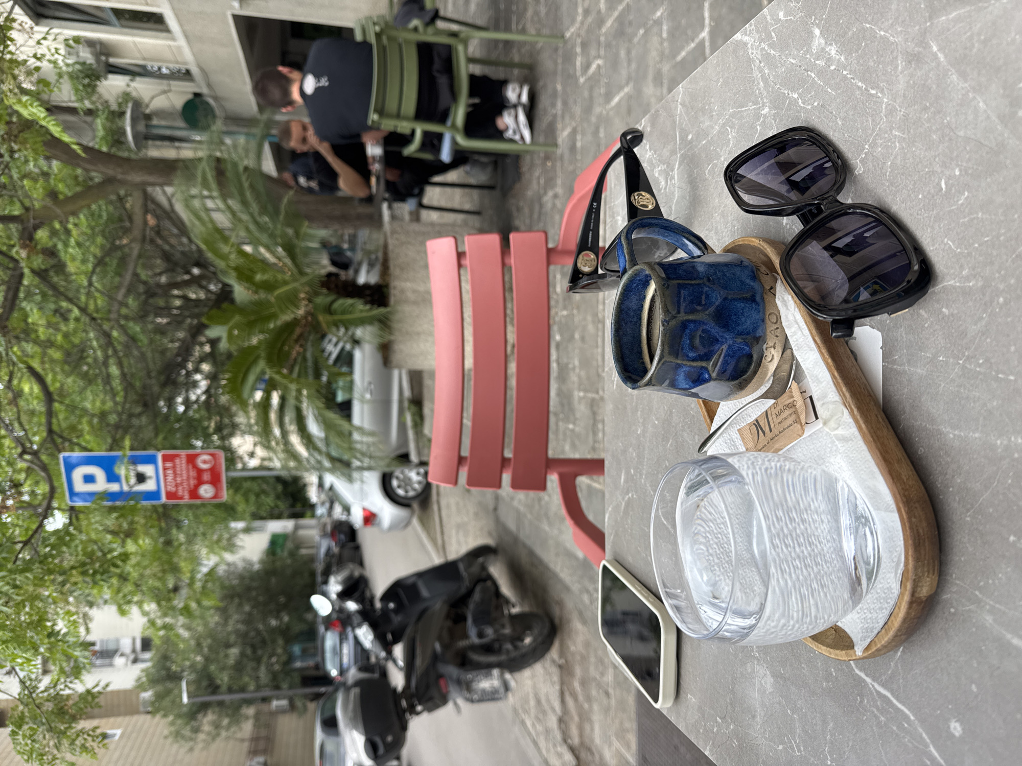 The image depicts an outdoor cafe setting, likely in Budva, Montenegro, given the restaurant coaster on the tray. A table with a gray marble-like surface holds a wooden tray containing a glass of water, a blue ceramic mug, and a pair of sunglasses. Two sets of sunglasses sit on the table. Beside the tray is a white-cased smartphone. A pink metal chair sits next to the table. In the background, other cafe patrons sit at green metal chairs and tables. Street parking signs are visible across the street, where several motorcycles and cars are parked. Trees line the street, and buildings are visible in the background.