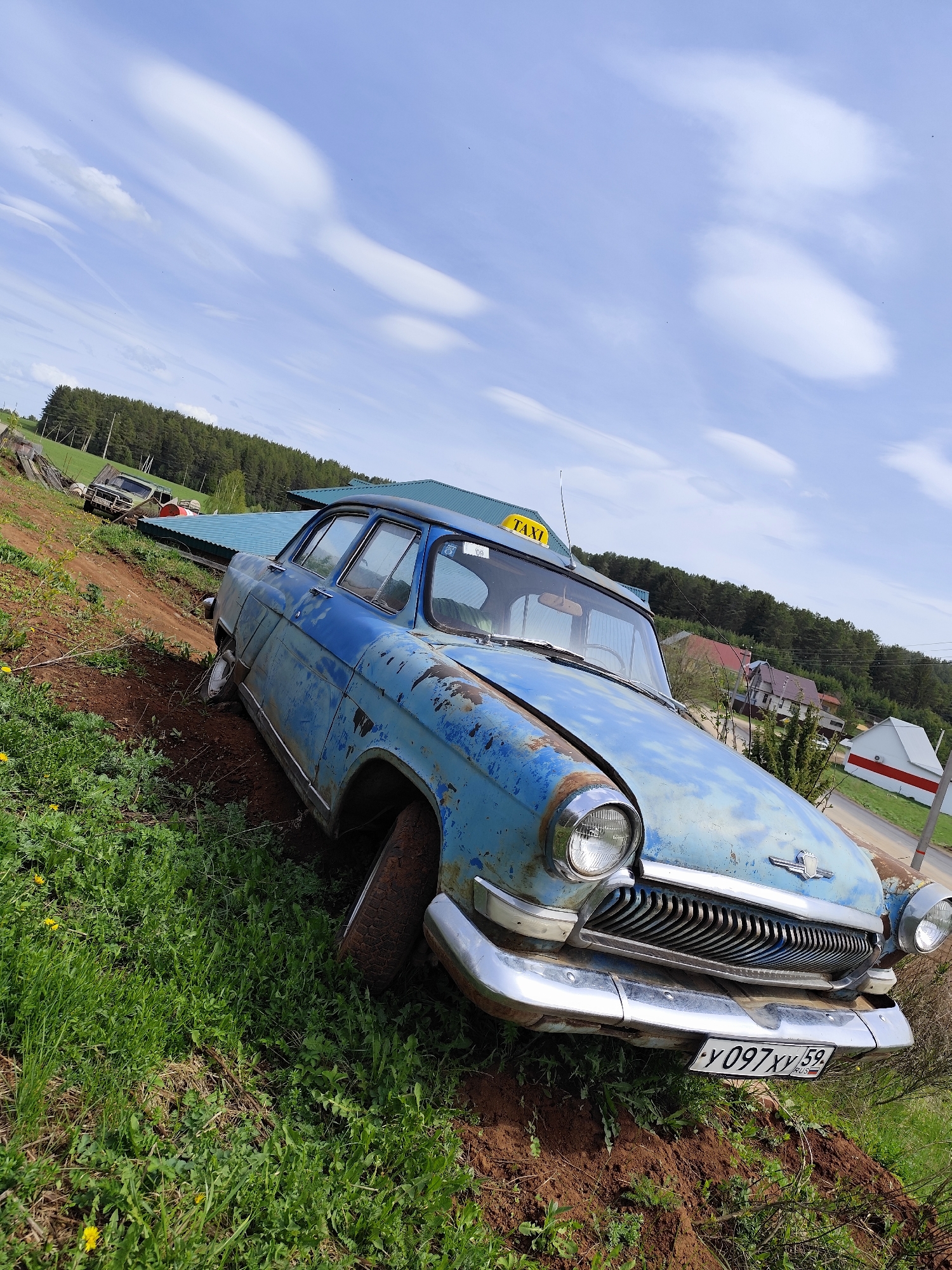 An abandoned, blue, vintage GAZ-21 Volga taxi is parked on a grassy hill in Фоки, Russia. The car exhibits rust and deterioration. It has a chrome bumper and a taxi sign on the roof. The license plate reads "Y097XY 59 RUS". In the background, there is a forested area, buildings, and a rural landscape under a blue sky with scattered clouds. The car is partially embedded in the soil, suggesting it has been stationary for a significant period.