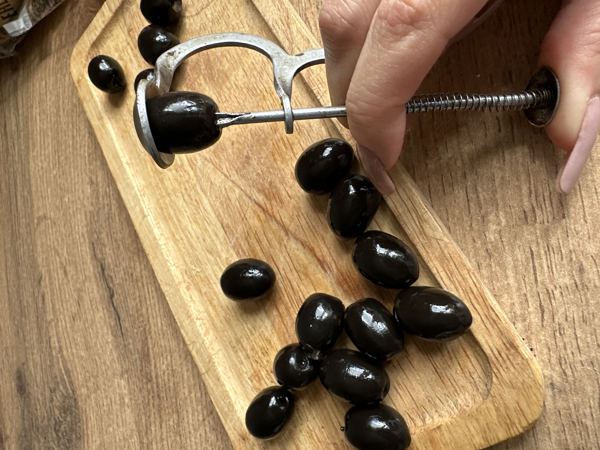 This overhead shot depicts a person in Stavropol, Russia, engaged in the activity of pitting black olives. A light-skinned hand, with long nails painted a light pinkish-nude color, holds and operates a metal olive pitter. One shiny black olive is secured within the pitter, indicating the pit removal process is underway.
The pitter itself appears to be a manual, spring-loaded type, showing signs of use. Numerous other shiny black olives are scattered across a light-toned wooden cutting board, which rests on a darker wooden surface, likely a table or countertop in a kitchen setting. Some olives on the board appear whole, while others may have already been processed. In the upper left corner, a blurry piece of packaging is visible, but any text on it is indiscernible. The image focuses on the practical task of preparing olives, highlighting the texture of the olives and the aged look of the kitchen tool.