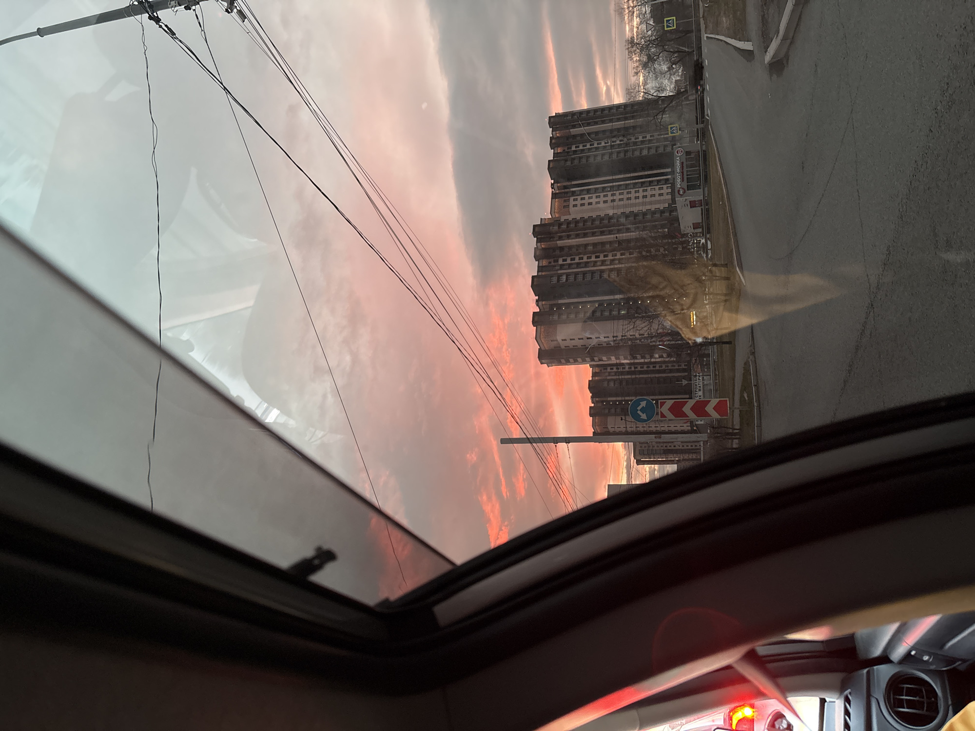 The view from inside a car through an open sunroof shows a street scene in Krasnoyarsk, Russia. A multi-story building is seen at the edge of the road. Traffic signs indicate the direction of travel. The sky is filled with orange and grey clouds. Power lines run overhead. The car's interior is visible in the foreground.