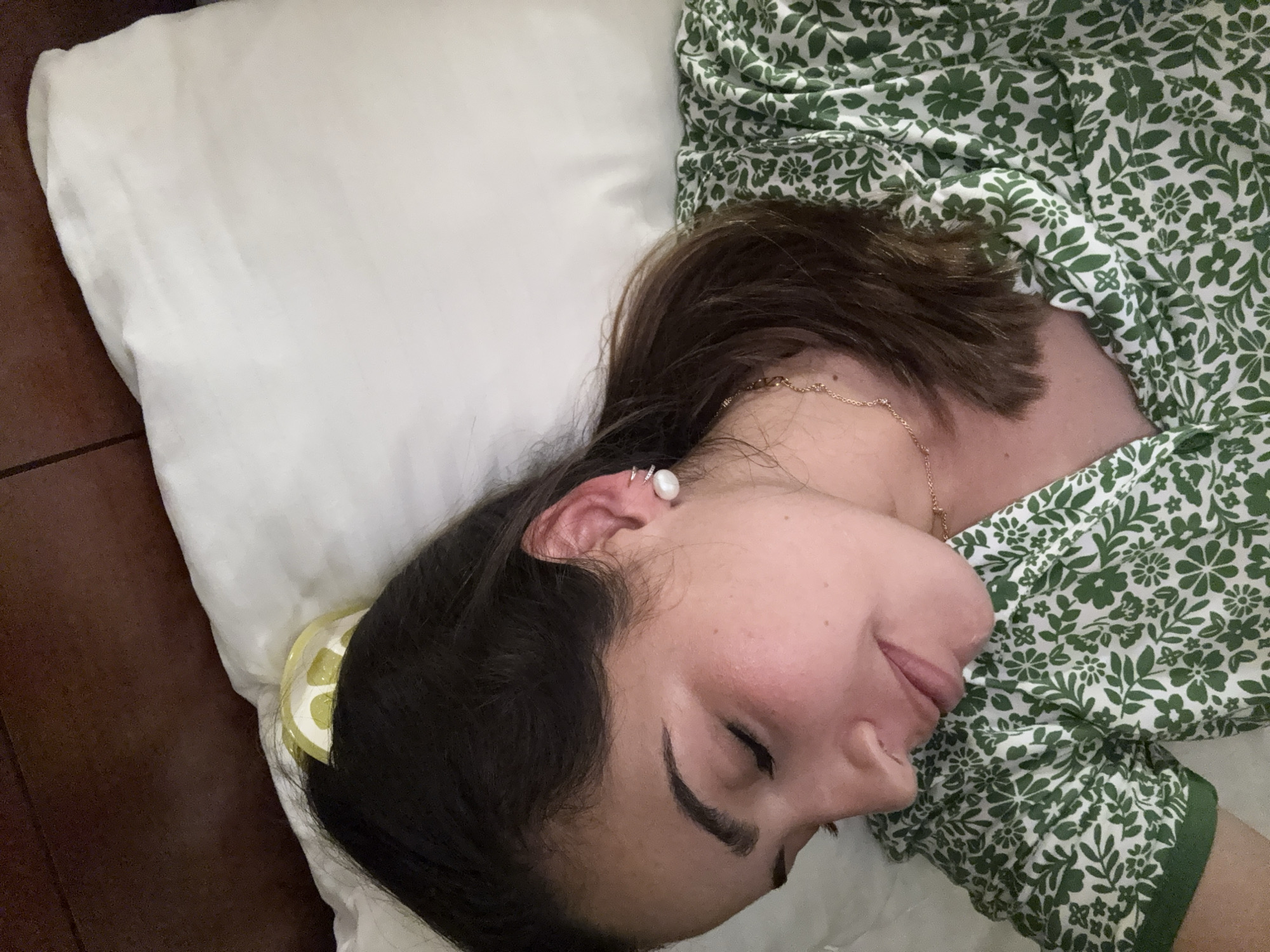 A woman is sleeping on a white pillow with a green and white floral dress. She has dark hair and is wearing an earring. A lemon-shaped hair clip is visible near her head. The location appears to be indoors, based on the lighting and surroundings. There is no information to confirm that the image was taken in San Antonio, United States of America.