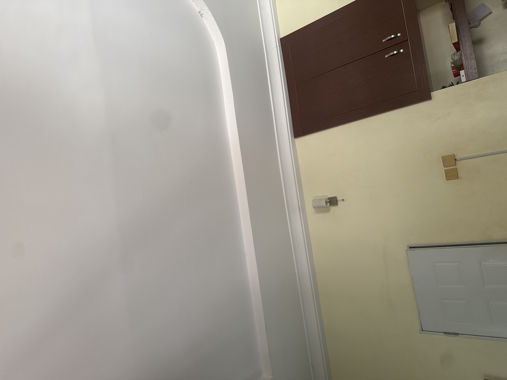 The image depicts an interior scene, likely a room, with a pale yellow wall and a white ceiling. A white door with four rectangular panels is visible on the left side of the frame. Above the door, a wall-mounted light fixture with a cylindrical shade is present. To the right of the door and light fixture, a dark brown cabinet with two doors and silver handles is mounted on the wall. Below this cabinet, a shelf is visible, containing various objects including what appears to be decorative items, a book, and a red cylindrical object. On the wall to the left of the cabinet, two light switches are installed. The room appears to be in a state of slight disarray, with some smudges and marks on the walls and ceiling. This scene is located in Narayi, Nigeria.