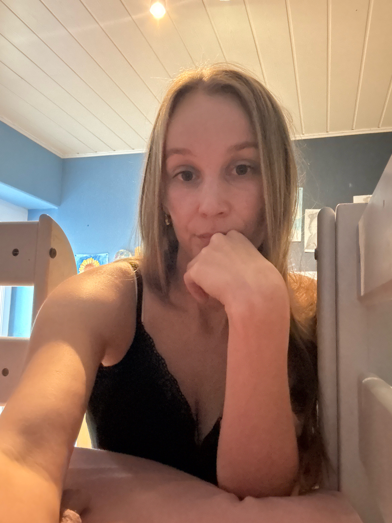 The image is a selfie taken indoors, likely in a bedroom. A young woman with long blonde hair is seated and looking towards the camera. She is wearing a black camisole with lace trim. Her right hand is resting on her chin, and her left arm is extended towards the camera, partially obscuring the view. In the background, a blue wall is visible with some artwork, and a white slatted ceiling with a light fixture. To the right and left of the woman are parts of a light-colored bed frame or bunk bed. There is no indication of the image being located in Überherrn, Germany.