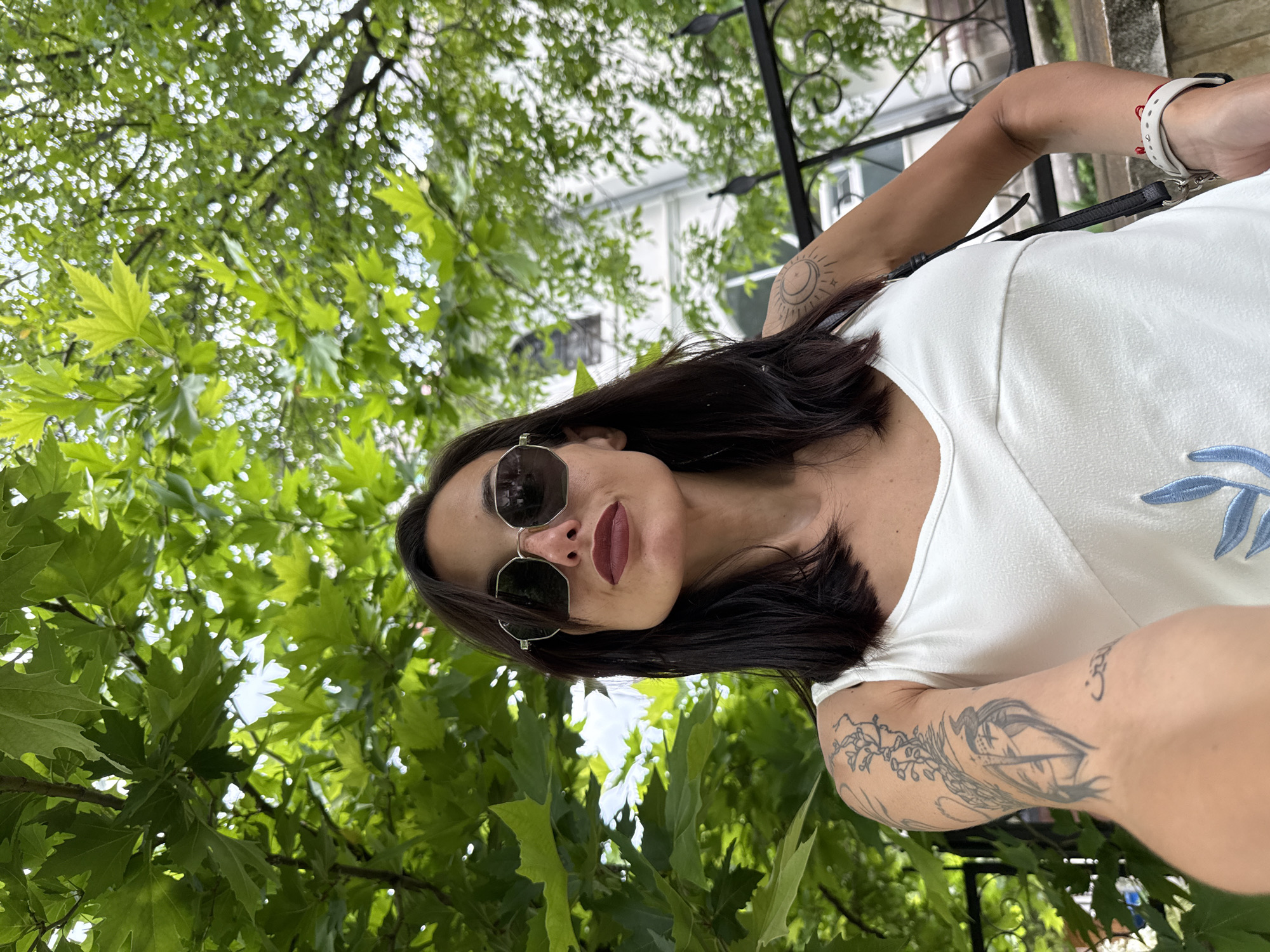 The image is a selfie taken outdoors, likely in a park or garden area of the city of Pleven, Bulgaria, as indicated by the foliage and architectural elements in the background. A woman with dark hair, wearing sunglasses and a white tank top, is the main subject. She has visible tattoos on her left arm and possibly her right shoulder, which include intricate designs. In the background, lush green leaves from trees dominate the upper portion of the frame, with a white building and a black metal fence visible further in the distance. The woman is looking directly at the camera, and her pose suggests she is standing. The lighting indicates it is daytime, possibly overcast or with diffused sunlight filtering through the leaves.