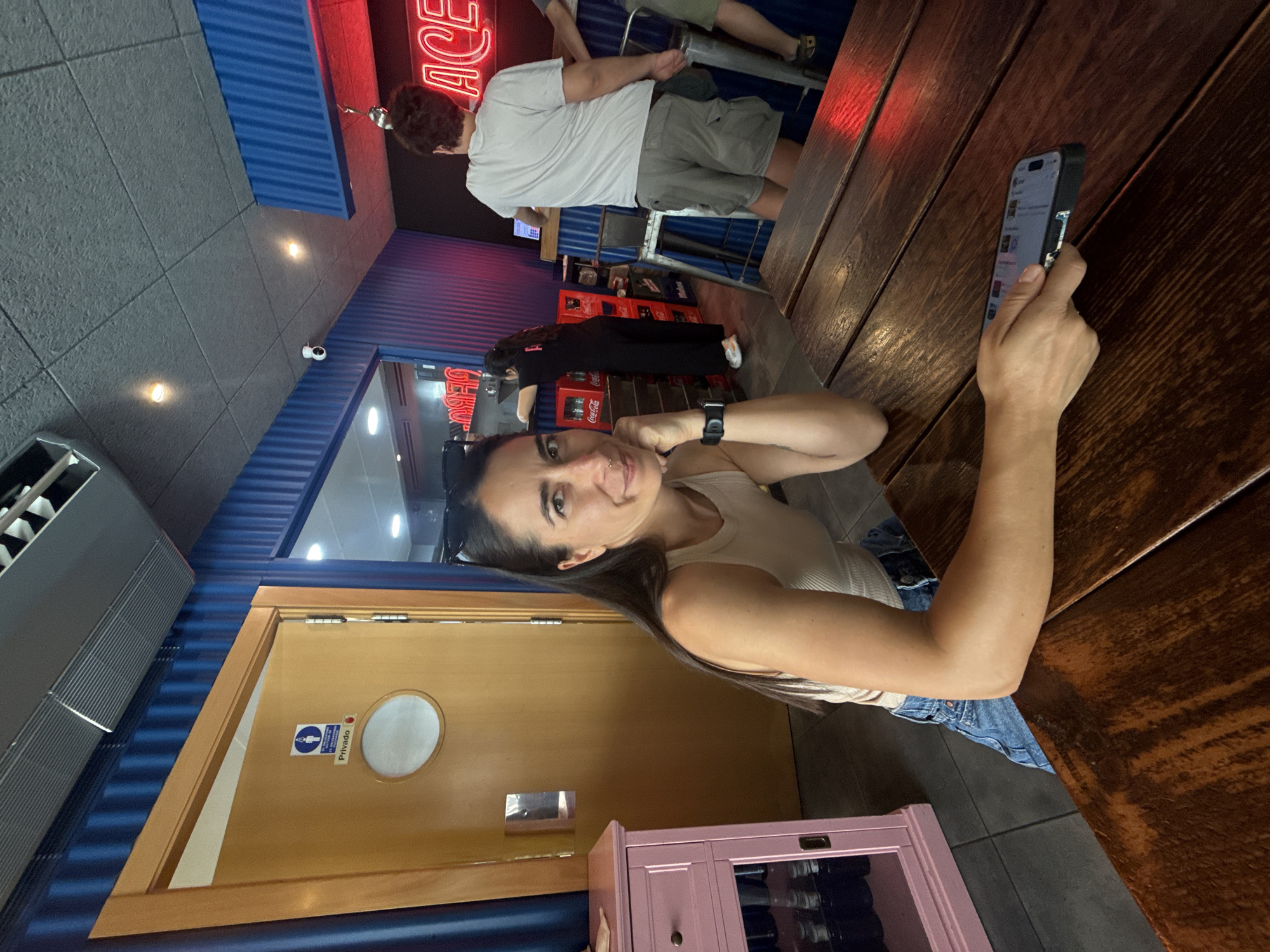 A woman with long dark hair and a nose ring, wearing a beige tank top and denim shorts, sits at a wooden table in a restaurant. She is looking at the camera and resting her chin on her hand. Her left arm is on the table, and she is holding a smartphone. In the background, a man in a white t-shirt and grey shorts stands near a neon sign that says "ACE". Another person, wearing a black outfit, is visible behind a counter. The restaurant walls are painted blue, and there's a wooden door with a "Privado" sign and a circular window. A pink cabinet is to the left of the door. The scene appears to be in Barcelona, Spain, given the "Privado" sign, a Spanish word for "private."