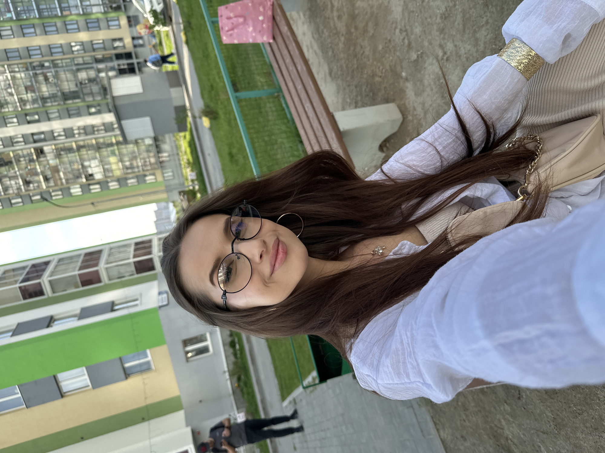 A woman with long brown hair, wearing round glasses, large hoop earrings, and a white blouse with a gold bracelet, is sitting on a wooden bench in an outdoor urban setting. She has a light-colored handbag with a gold chain strap next to her. A tall apartment building with green, grey, and brown panels is visible in the background. Two men are walking on a paved area near the building. The location is likely in a residential area of Yekaterinburg, Russia, based on the architectural style of the apartment building.