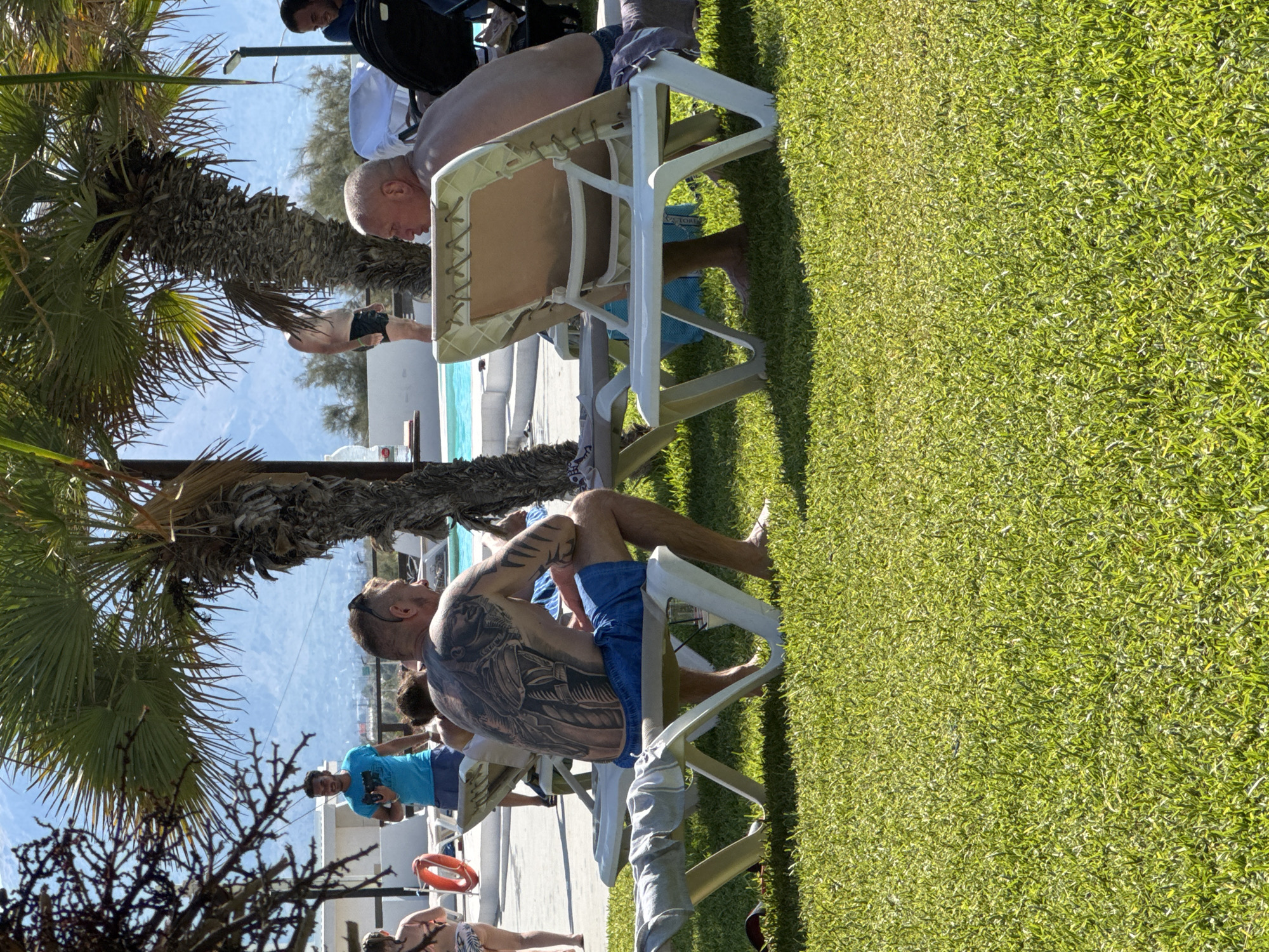 In Isola delle Femmine, Italy, two men are seated on lounge chairs on a grassy area near a swimming pool. The man on the left, facing away from the camera, has a large, intricate tattoo covering his back and is wearing blue shorts. The man on the right, facing to his left, is bald and is also wearing shorts. Several other people and palm trees are visible in the background, along with lounge chairs and a swimming pool. The environment appears to be a sunny outdoor recreational area.