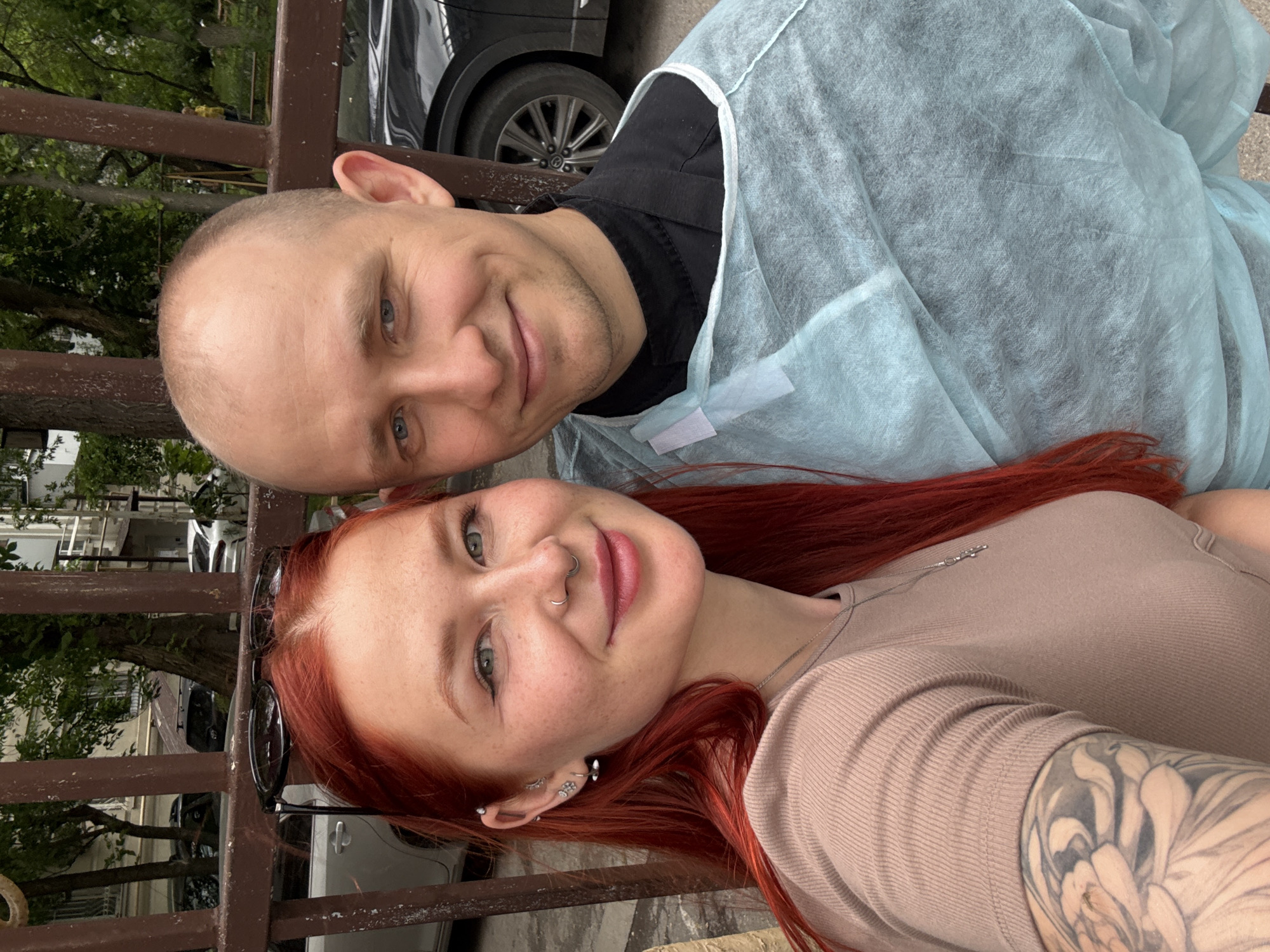 A selfie taken outdoors in an urban area shows a man and a woman. The woman is in the foreground, with red hair, a nose ring, multiple ear piercings, and a large tattoo on her arm. She wears a light brown ribbed shirt. Behind her and to the left is a partially visible brown metal fence. Behind the fence are trees and buildings, with several vehicles visible, suggesting an urban setting.
The man, partially covered with a light blue medical gown, stands behind the woman. He is bald with fair skin. A black t-shirt is visible under the medical gown. A portion of a black vehicle is visible in the background behind him. The photo's location is Rostov-on-Don, Russia.