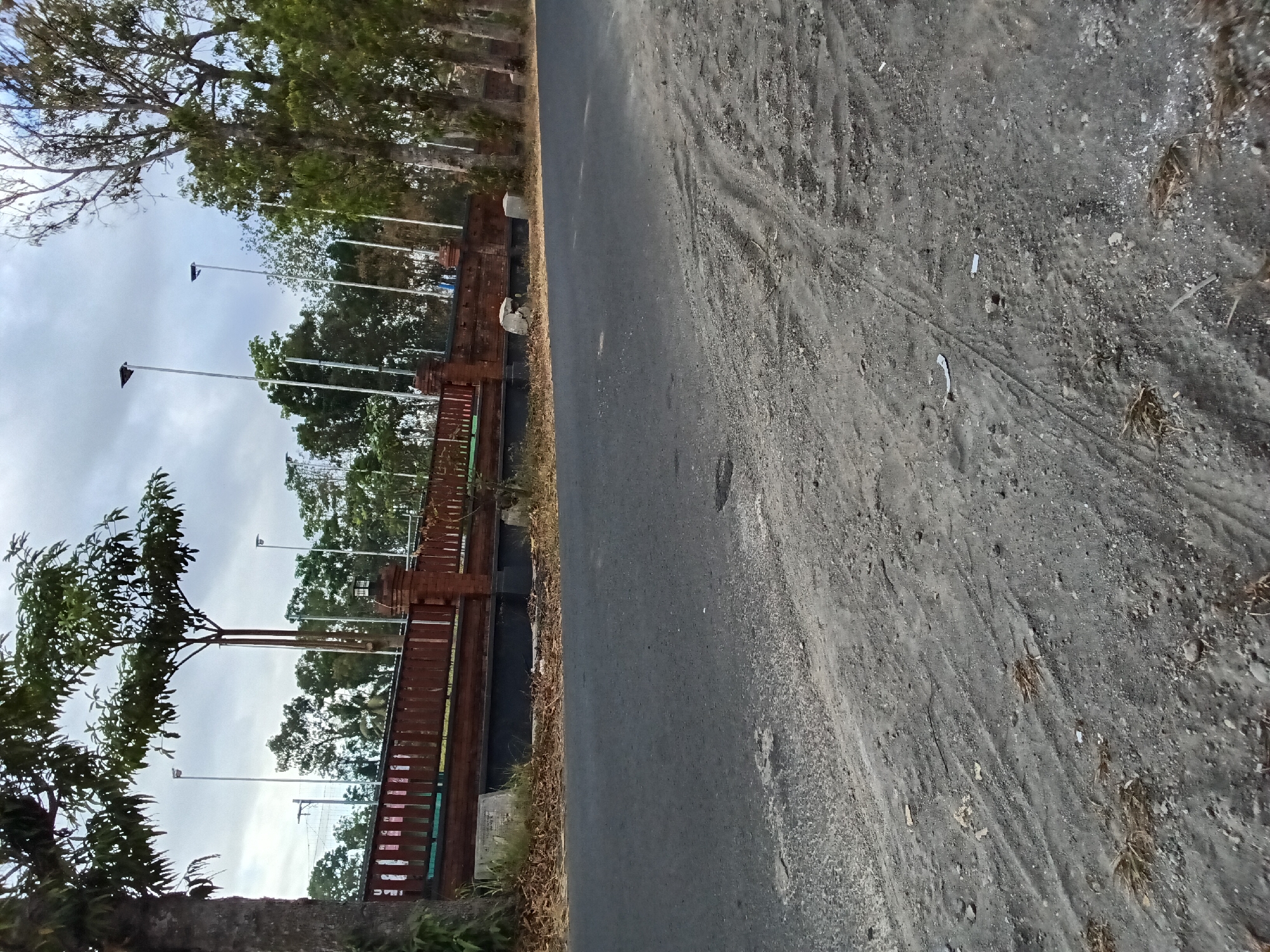 The image depicts a roadside scene in Bendorejo, Indonesia, on an overcast day. In the foreground, a paved asphalt road stretches towards the right, with a dusty, gravelly shoulder on the left, marked by tire tracks. A low, red brick wall with a wooden fence runs along the far side of the road, separating it from a line of trees and what appears to be a park or recreational area. Several tall, slender lampposts are visible behind the wall. Large trees, some with dense foliage and others with sparser branches, line the right side of the road. The ground is covered in dry leaves and debris. No people or vehicles are present in the image.