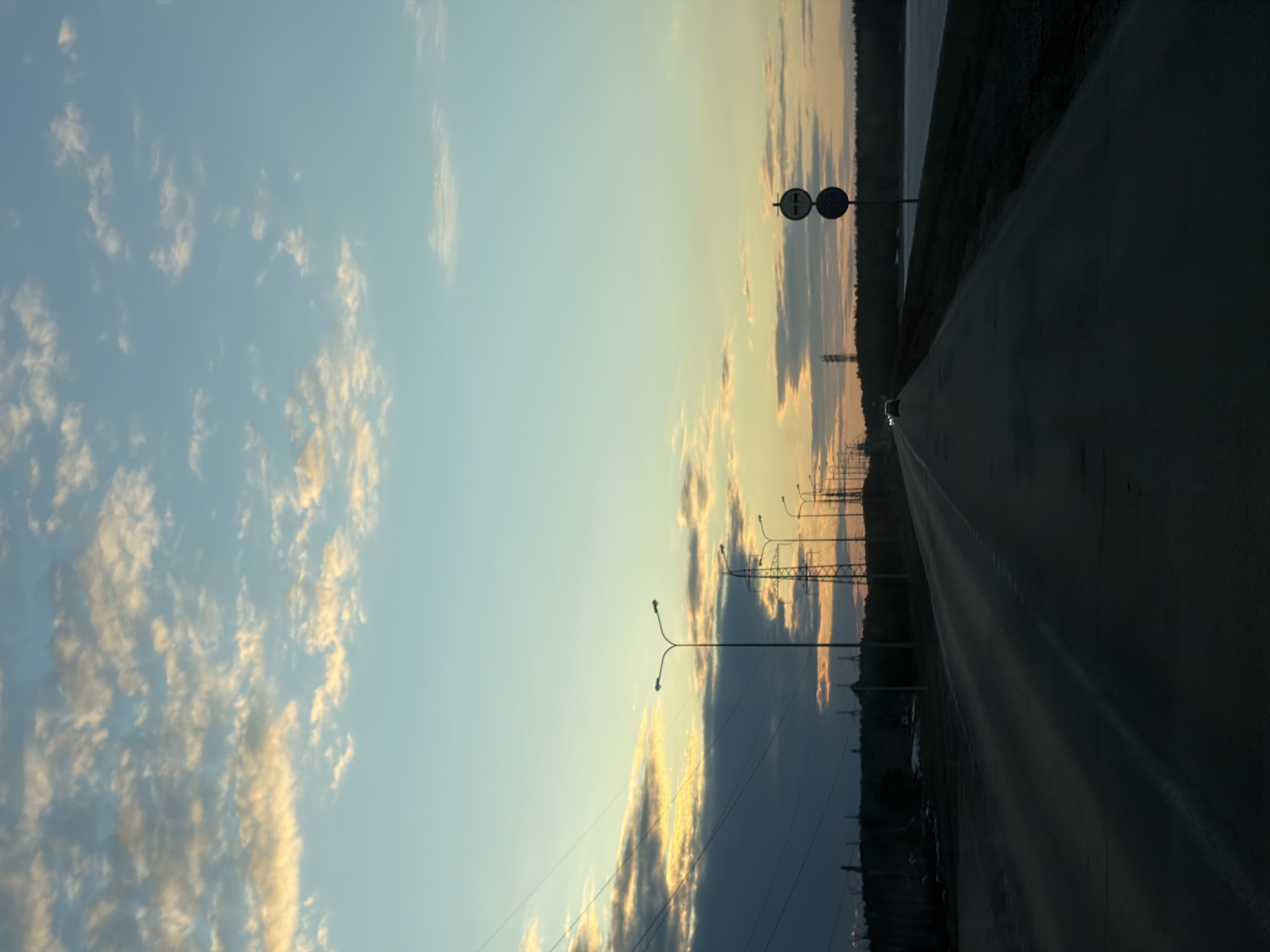 The image depicts an asphalt road stretching into the distance under a sky transitioning from blue to orange and yellow at sunset. On the left, the sky is filled with scattered clouds illuminated by the setting sun. Along the road, evenly spaced streetlights line the edge, extending toward a silhouetted treeline on the horizon. A road sign is visible. A single vehicle, its headlights visible, is present on the road in the distance. The location is Chaikovsky, Russia.