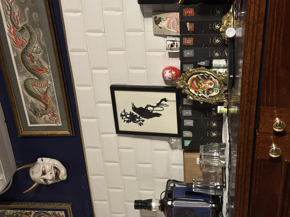 A narrow, vertical composition presents framed artwork, including a dragon, a black bird with flowers, and a mask on a blue wall. The right side features a stack of black boxes, a framed tattoo image, glasses, and a liquor bottle next to a wooden cabinet with round knobs. The background is a white tiled wall.