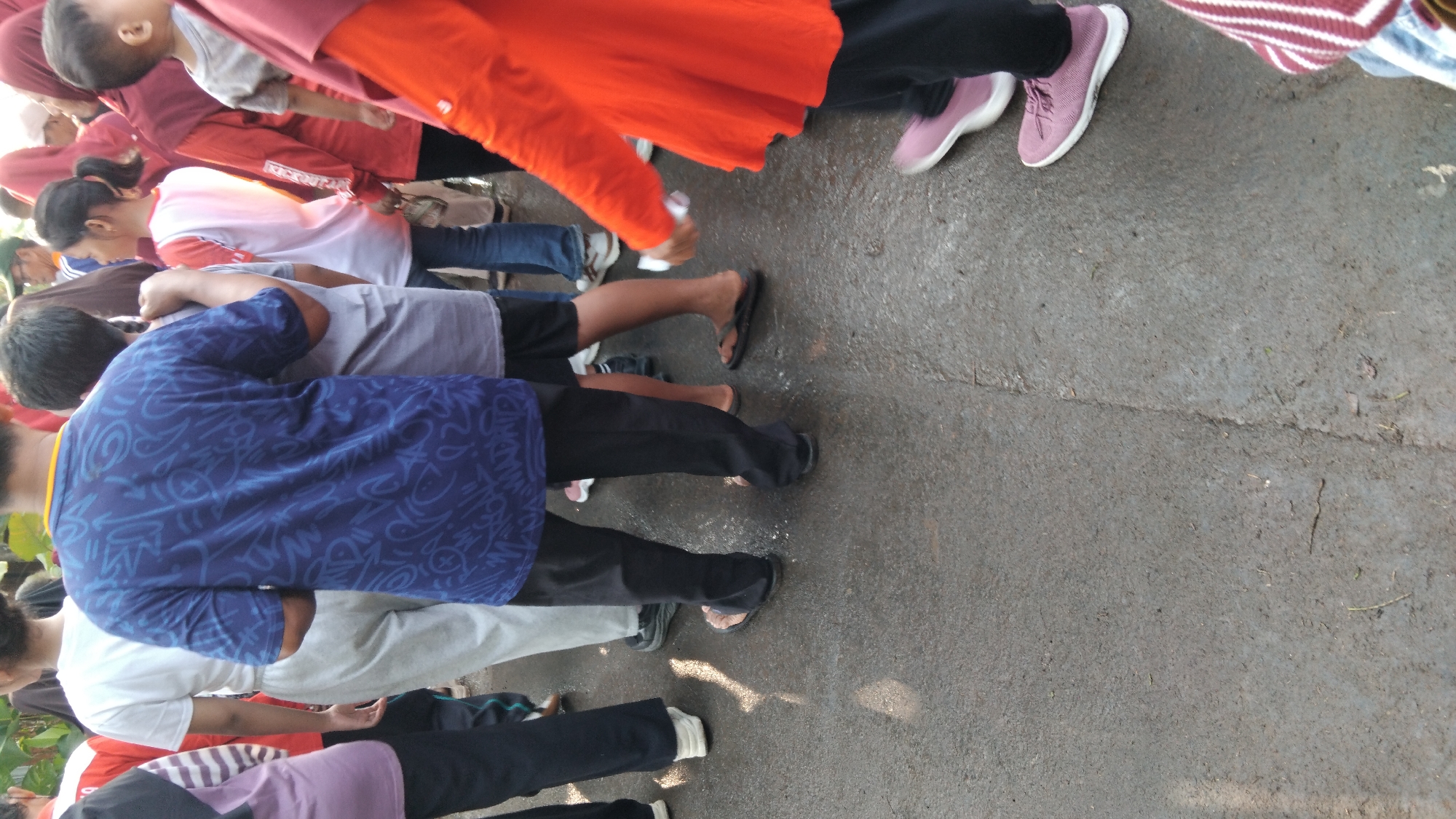 This is a medium shot of a crowd of people standing on a wet, asphalt surface in Posong, Indonesia. The perspective is from behind and slightly to the side, focusing on the lower bodies and midsections of the individuals. Several people are wearing casual clothing, including t-shirts, pants, and shorts. One person in the foreground is wearing a dark blue t-shirt with a white graphic design, black pants, and black sandals. Another individual to their left is wearing black pants and white shoes. To the right, a person in an orange long-sleeved shirt is visible. The ground appears to be wet and reflective, suggesting recent rain or a damp environment, common in the tropical climate of Posong. The overall scene depicts a gathering of people in an outdoor setting, possibly a street or public area, with a focus on their attire and the ground surface.