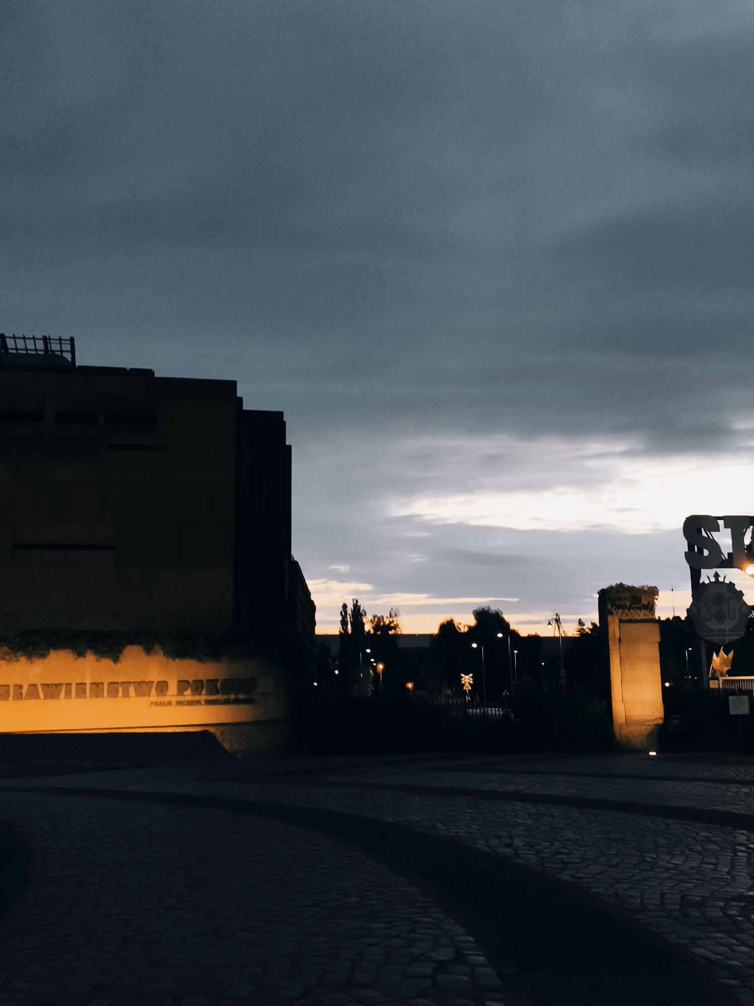 The image depicts a street scene at dusk in Gdańsk, Poland, likely near the Monument to the Fallen Shipyard Workers. The foreground features a cobblestone street with visible tram tracks. To the left, a large, dark building with some illuminated text on its facade is prominent. The text appears to be in Polish, possibly related to the shipyard or its history, with the words "SŁAWIEŃSTWO POKON" clearly visible and illuminated by an orange light. In the mid-ground, silhouetted trees and streetlights are visible. To the right, a monumental structure, possibly part of the monument complex, is lit by a warm yellow light. Above, a cloudy sky transitions from dark gray to lighter tones with hints of sunset colors. The overall atmosphere is somber and evocative.